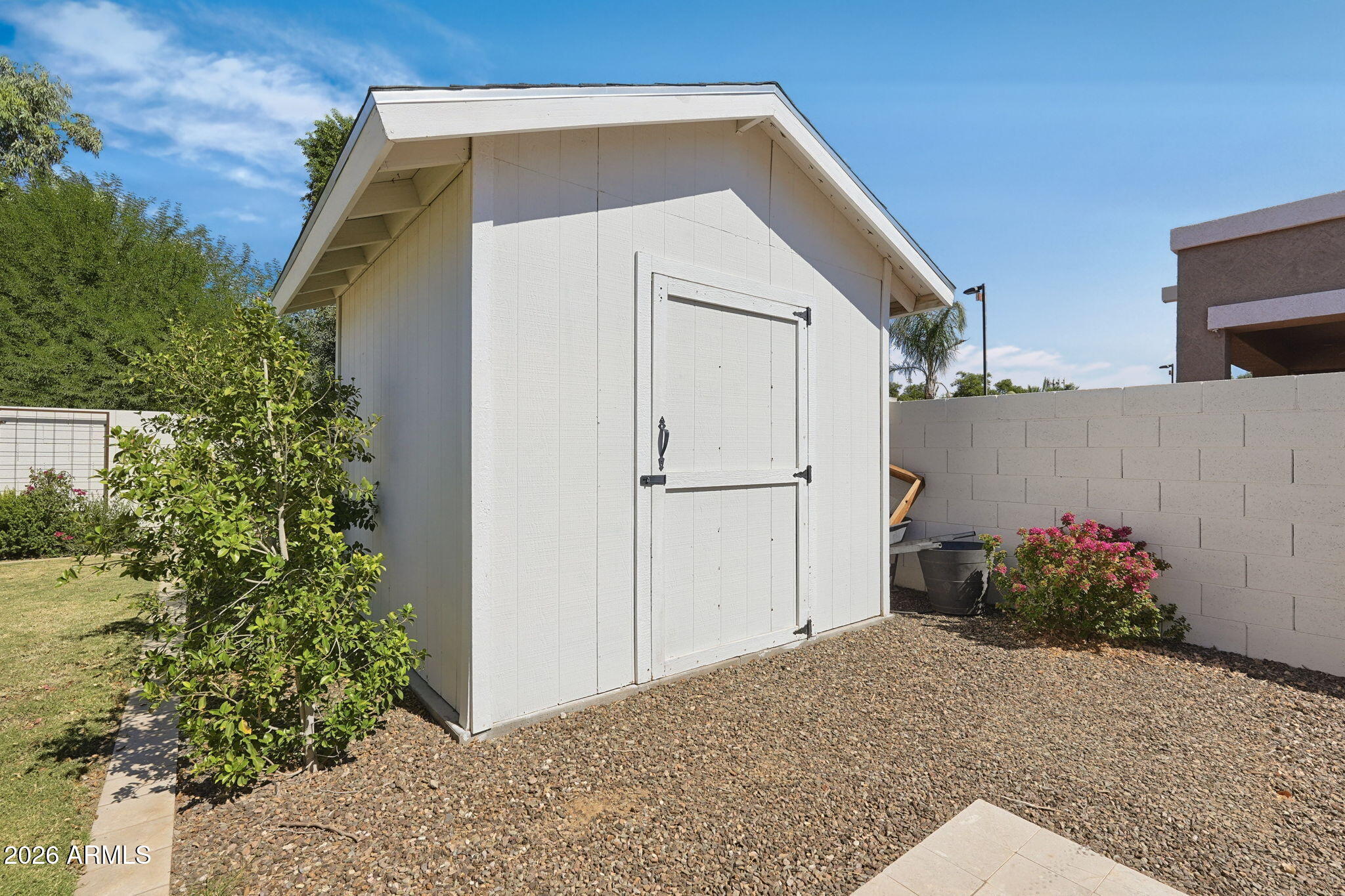 White backyard storage shed with double doors, gravel landscaping, shrubs, and a block wall.
