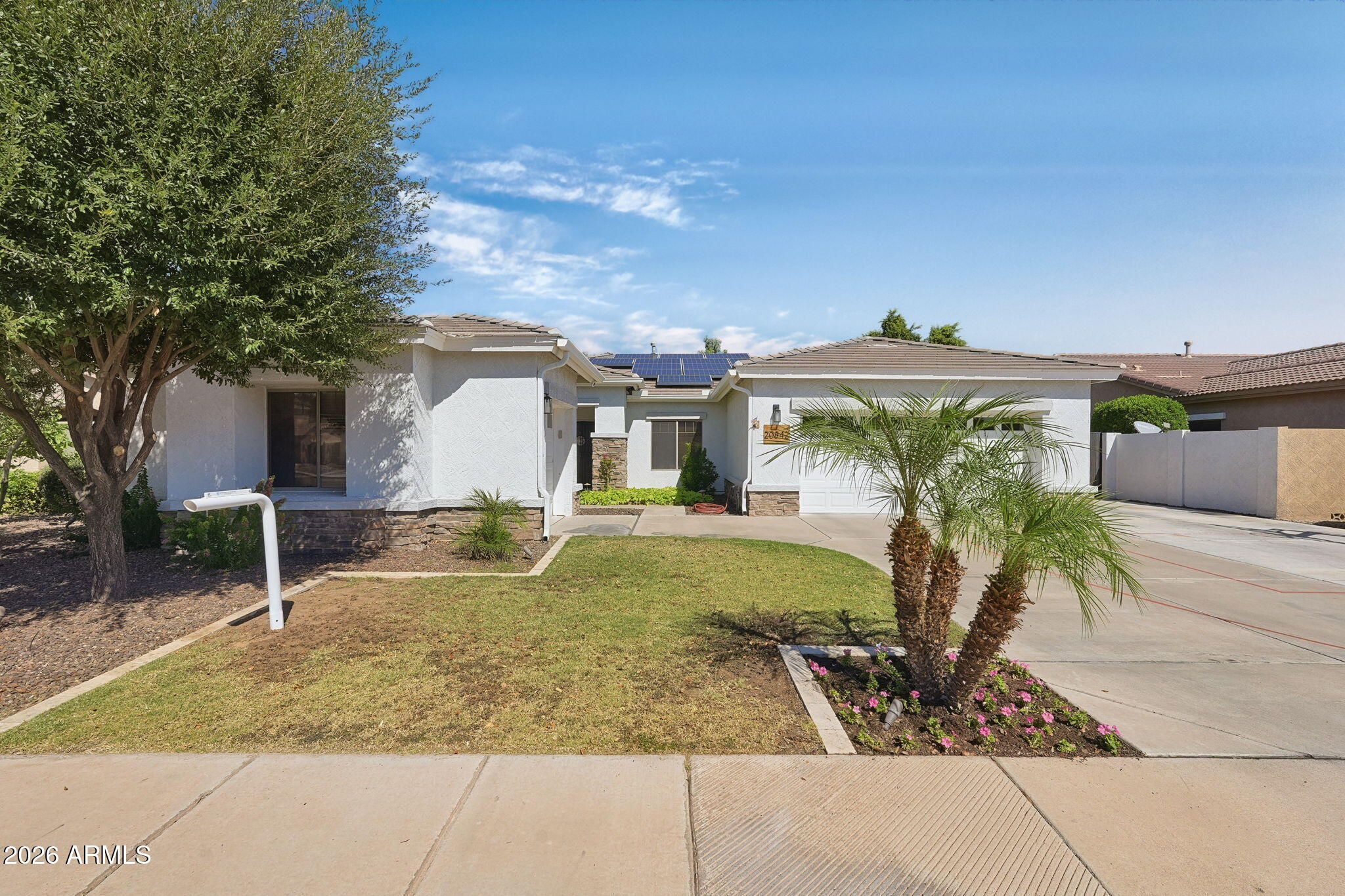 Single-story white stucco home with rooftop solar panels, a front lawn, and palm trees beside the driveway.
