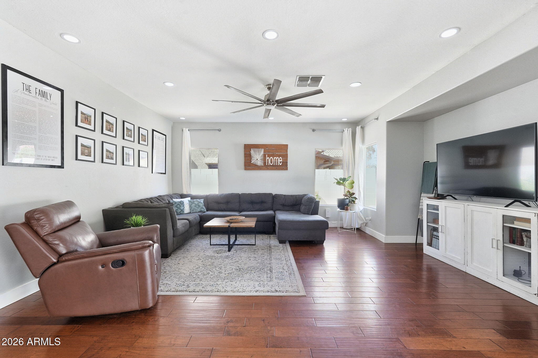 Living room with a gray sectional, leather recliner, ceiling fan, and reddish wood floors.