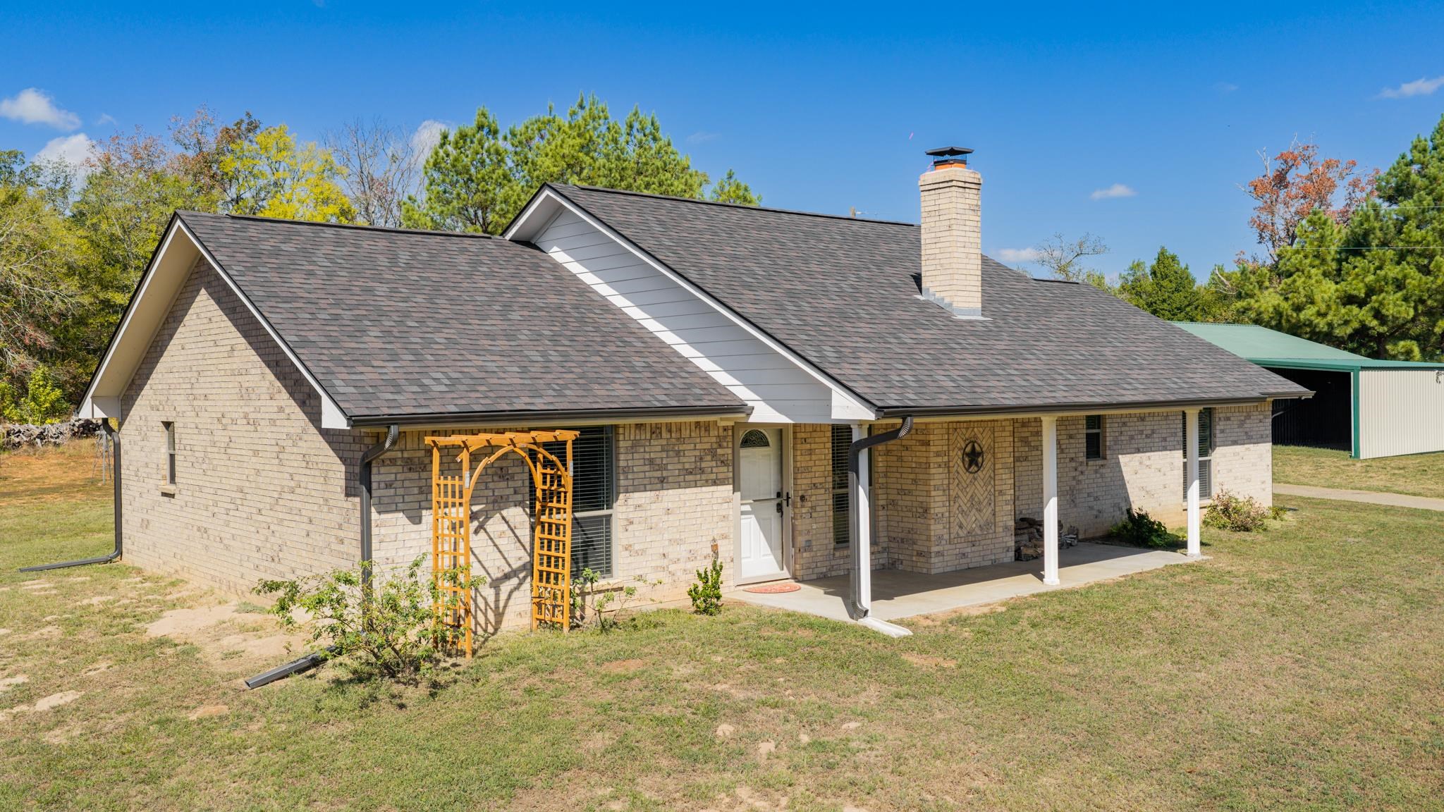 Light brick house with dark shingle roof, covered entry, and wooden garden arbor.