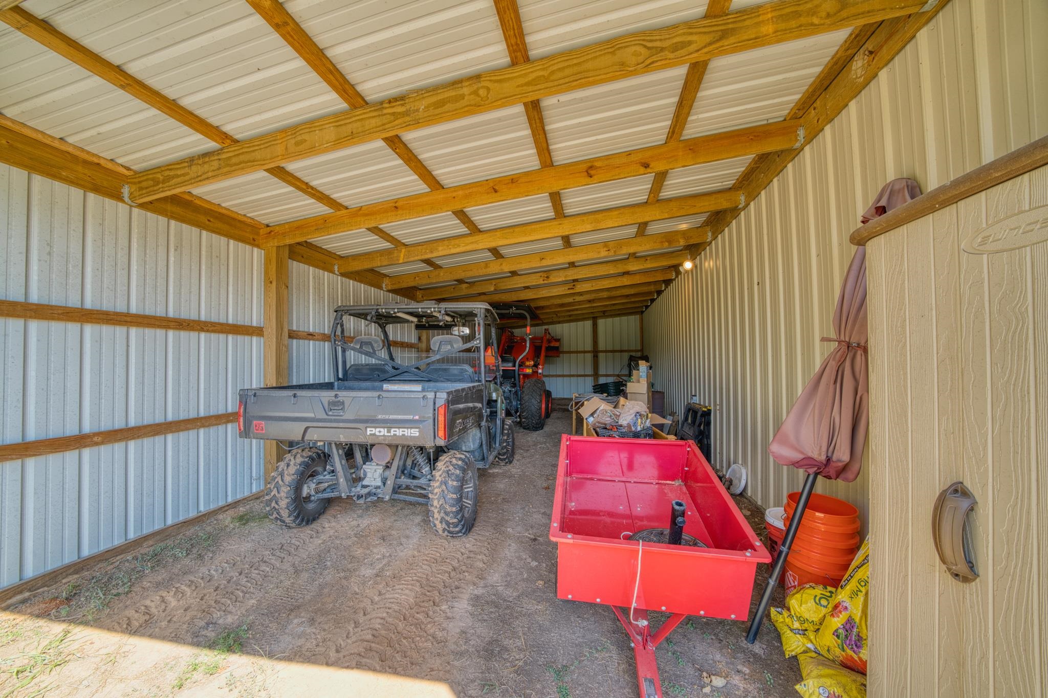 Storage shed with a Polaris utility vehicle, red trailer, and equipment beneath exposed wooden roof beams.