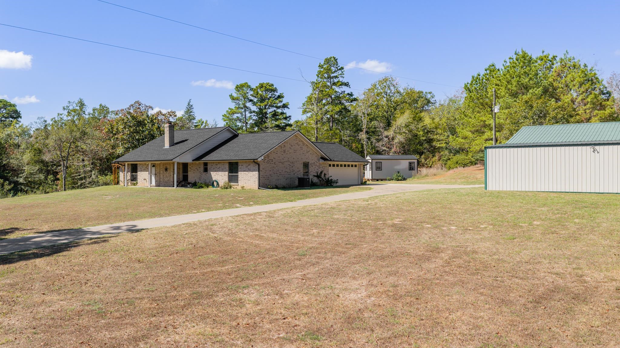 Brick ranch home with an attached garage, detached metal workshop, and small outbuilding on a broad lawn.