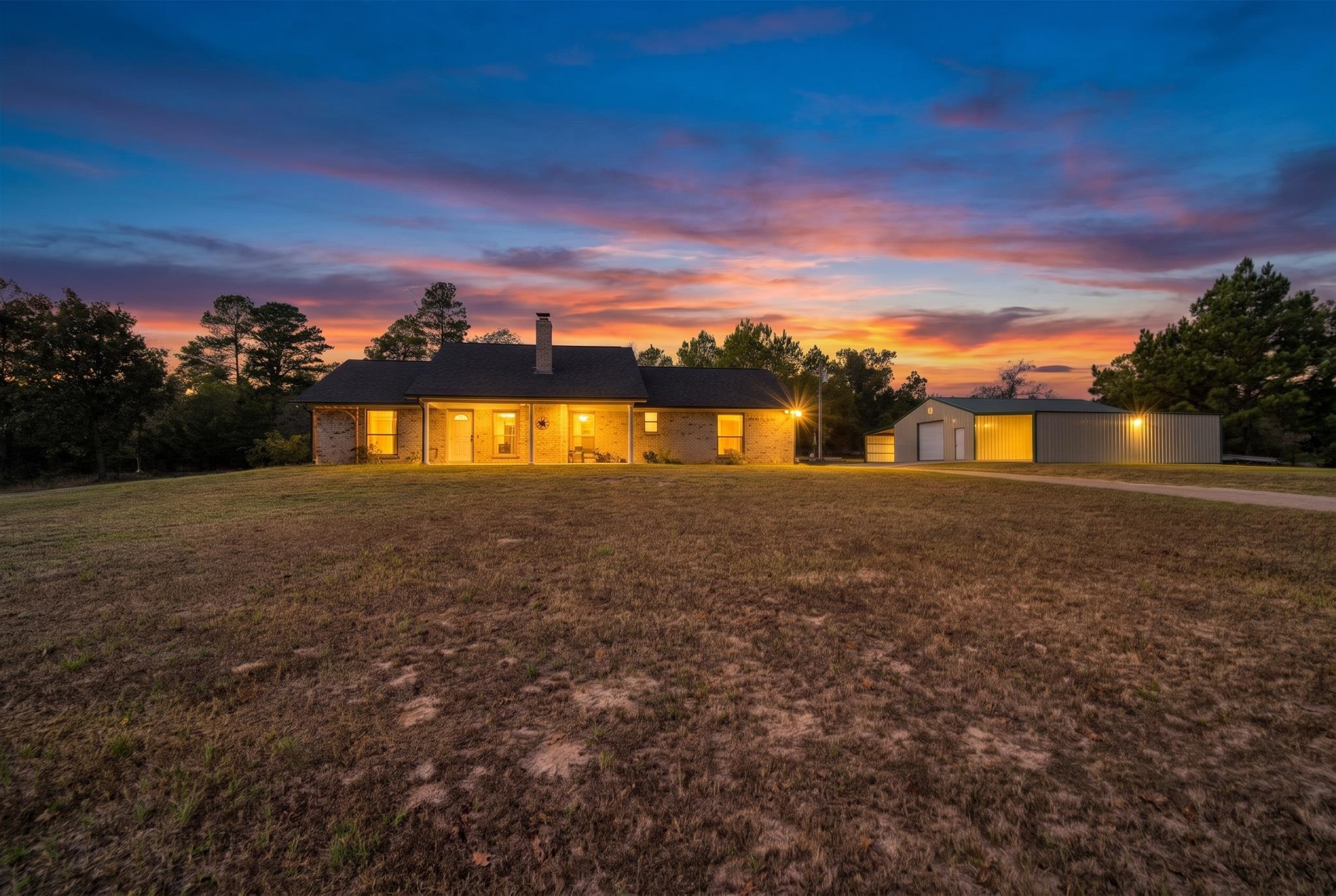 Brick house and metal outbuilding with warmly lit windows beneath a colorful sunset.