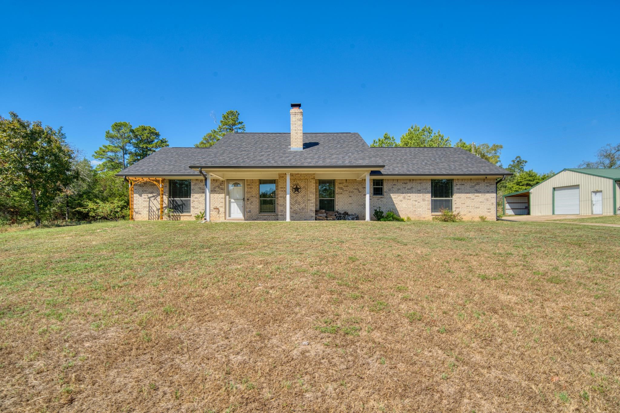 Single-story brick house with a covered porch, chimney, broad lawn, and detached metal outbuilding.