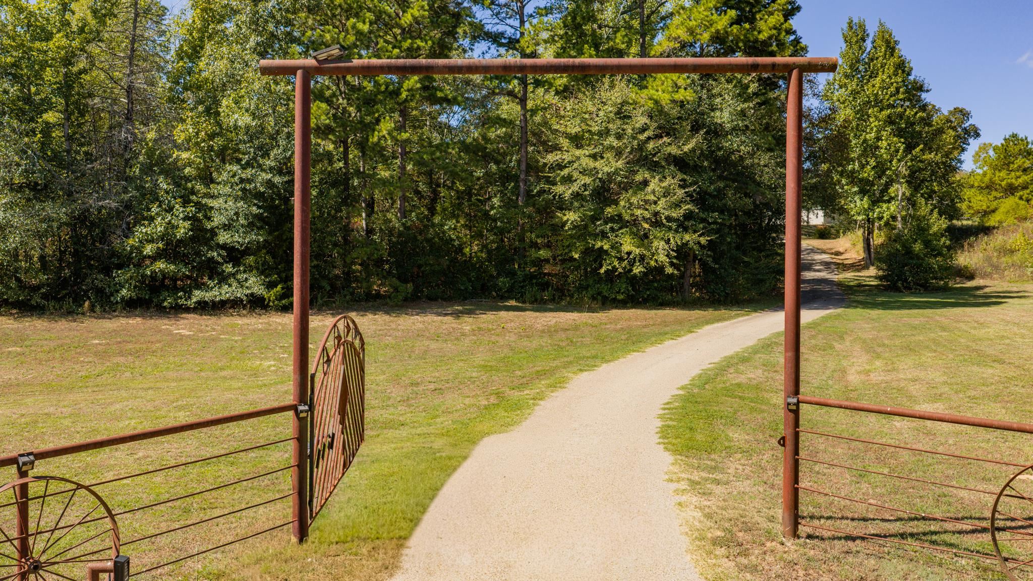 Gravel driveway through a metal gate, bordered by a grassy clearing and mature trees.