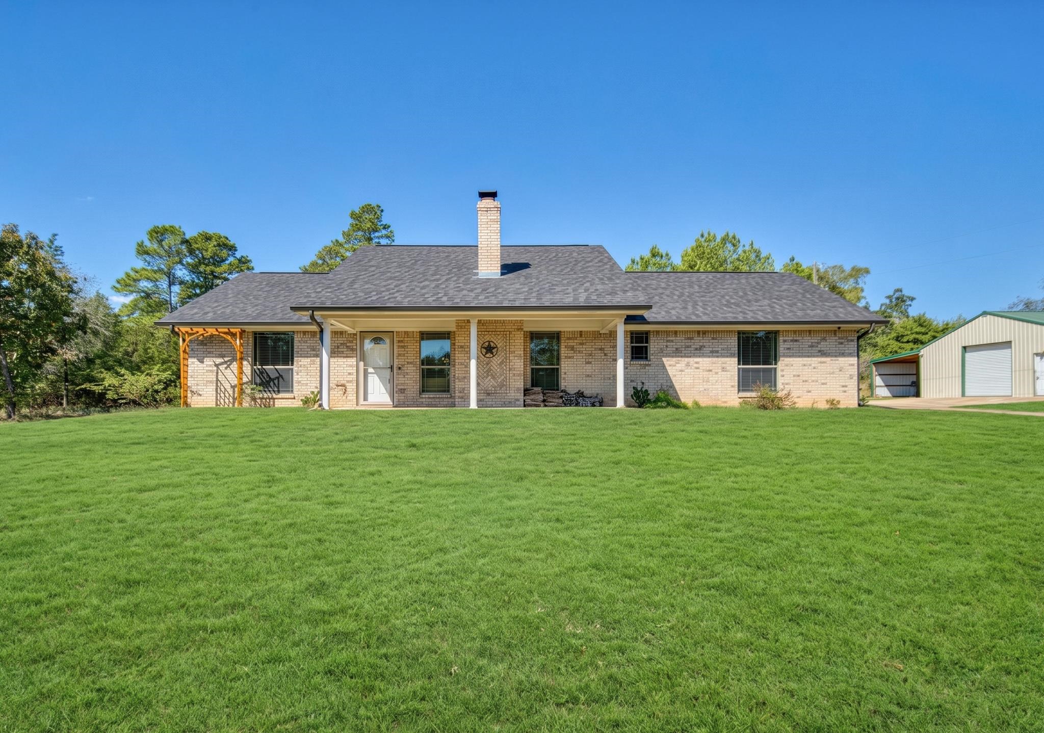 Single-story brick house with a covered porch, chimney, broad lawn, and detached metal outbuilding.