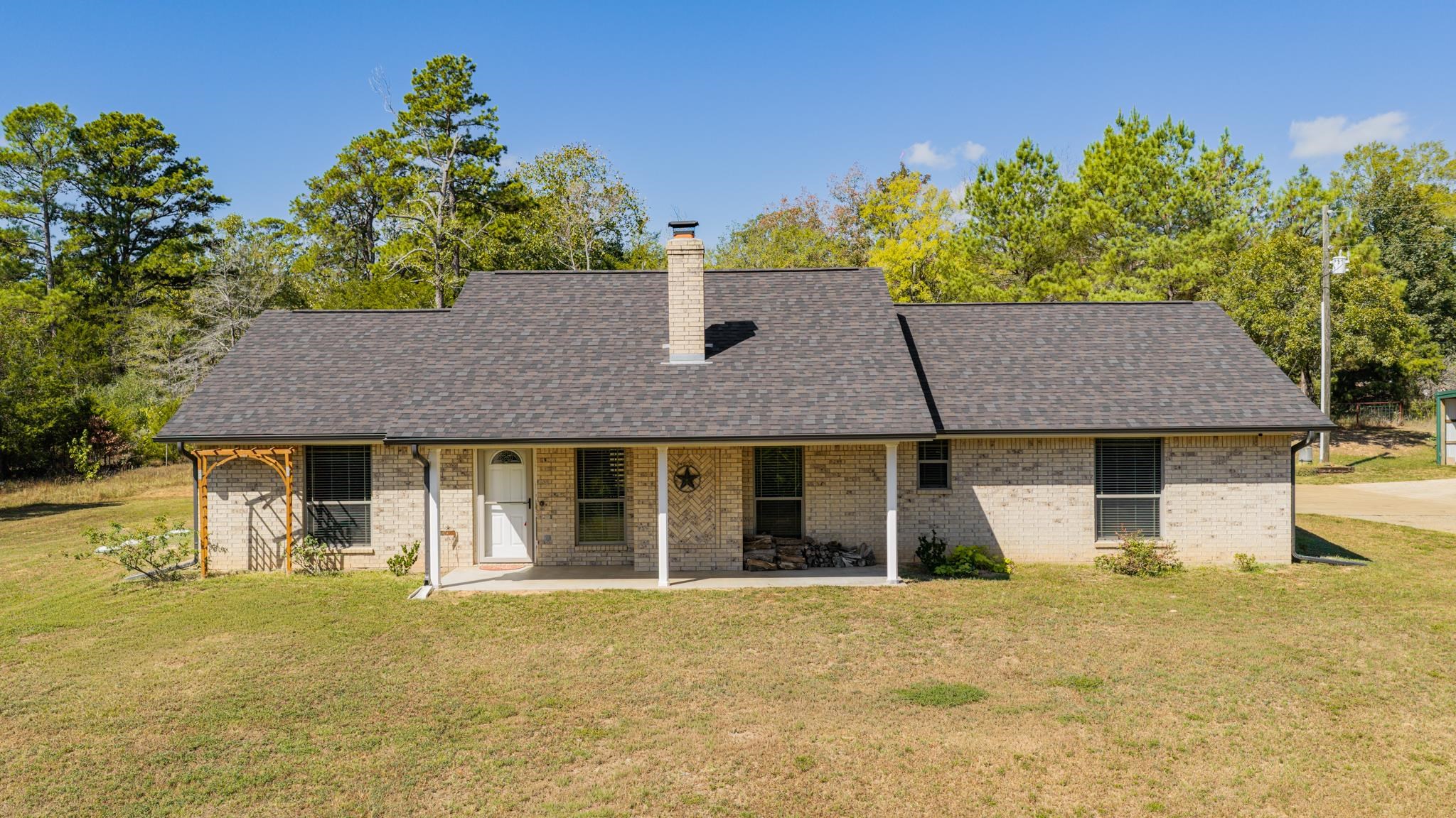 Single-story brick home with a dark roof, chimney, and covered porch, set against mature trees.
