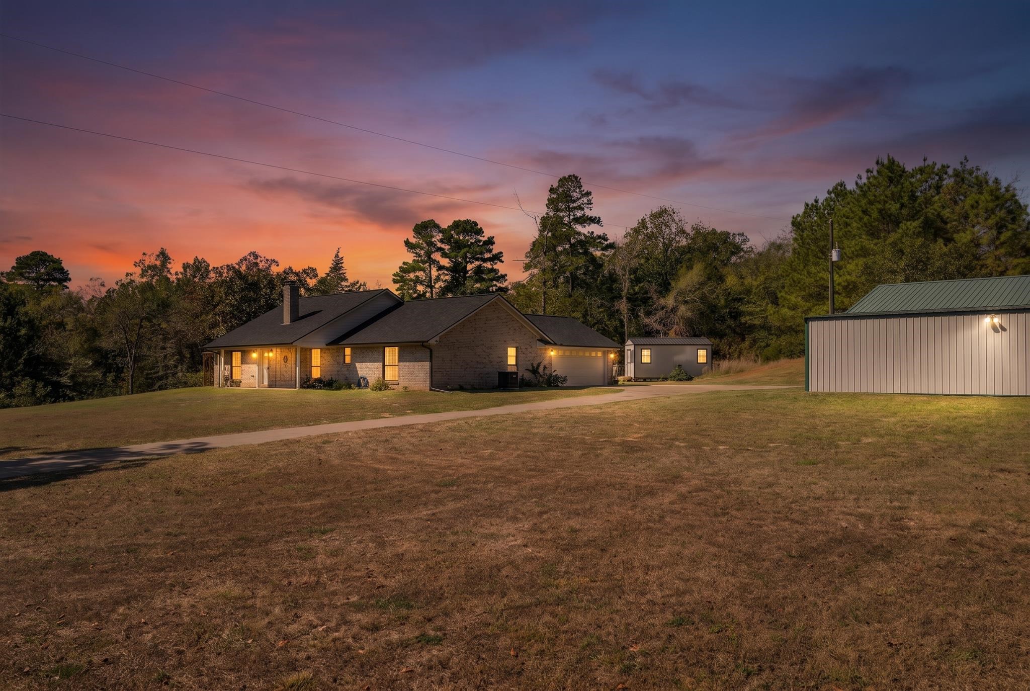 Brick house and detached metal building beside a broad lawn under a pink-orange dusk sky