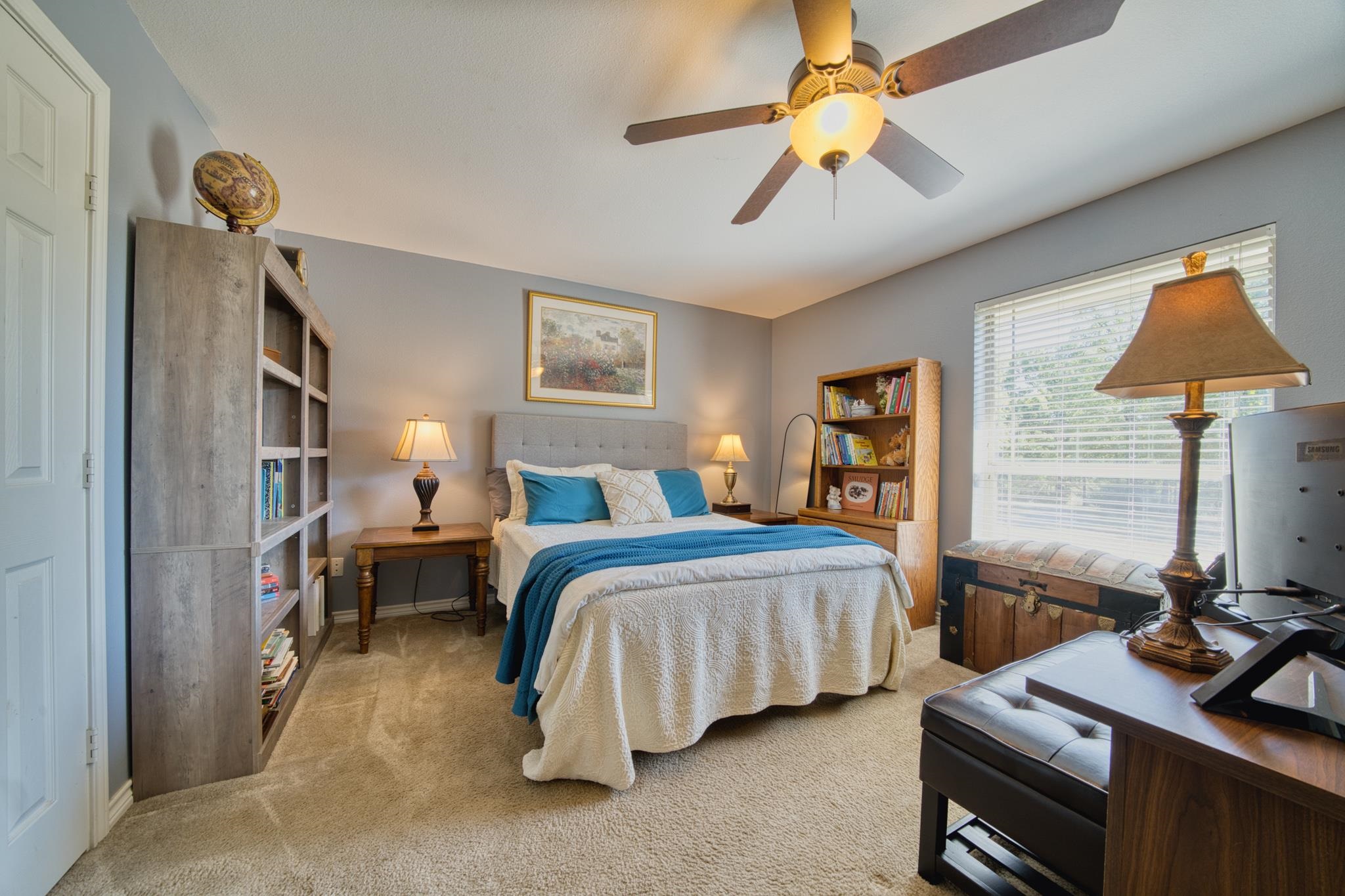 Furnished bedroom with a white-and-teal bed, bookshelves, ceiling fan, and large window.