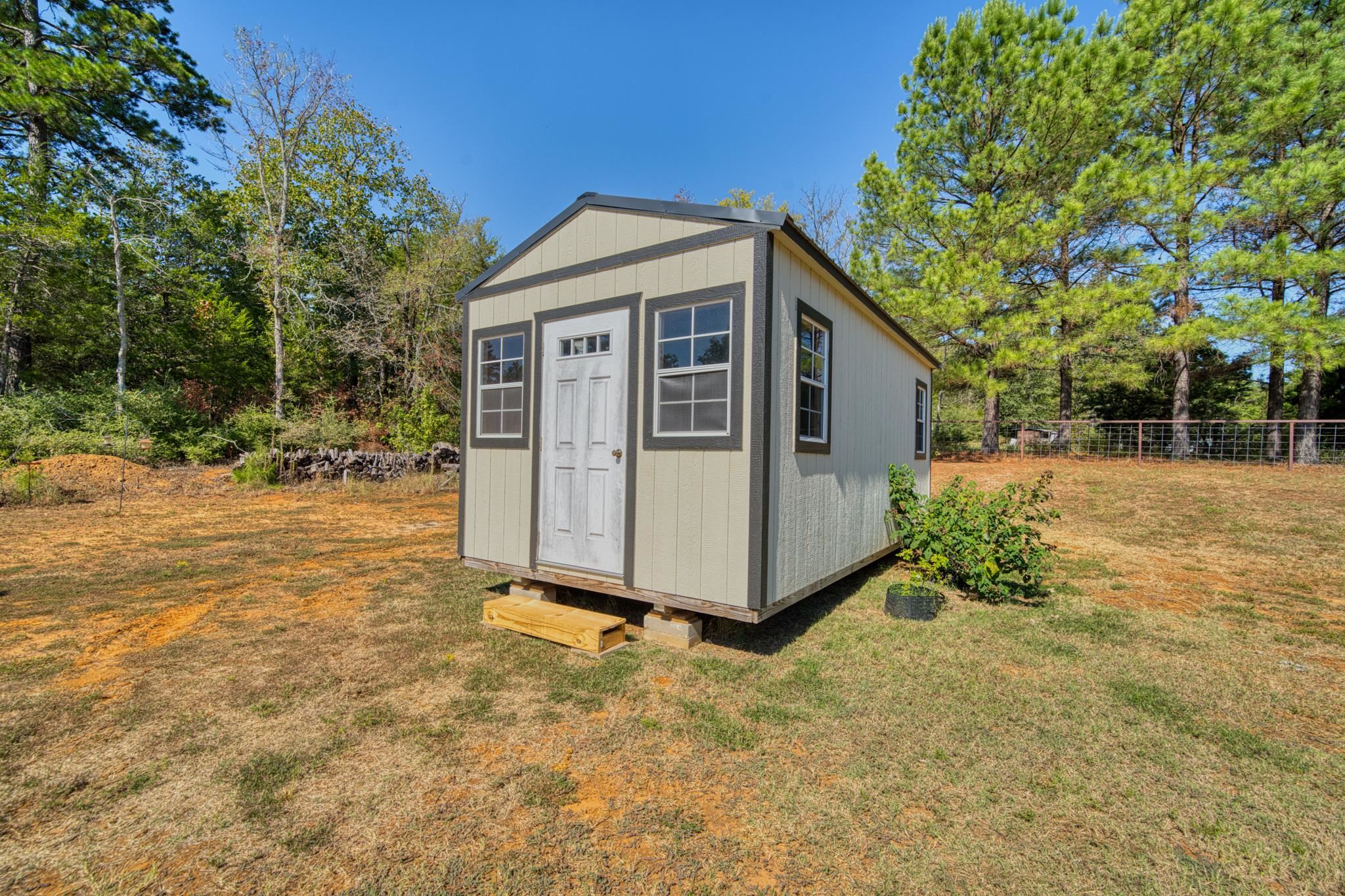 Beige detached shed with dark trim and a white door on a tree-lined lawn.