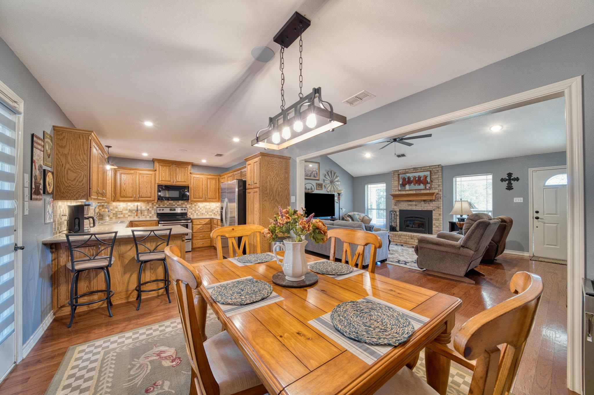 Open-plan kitchen and living room with wood cabinetry, a large dining table, and a brick fireplace.