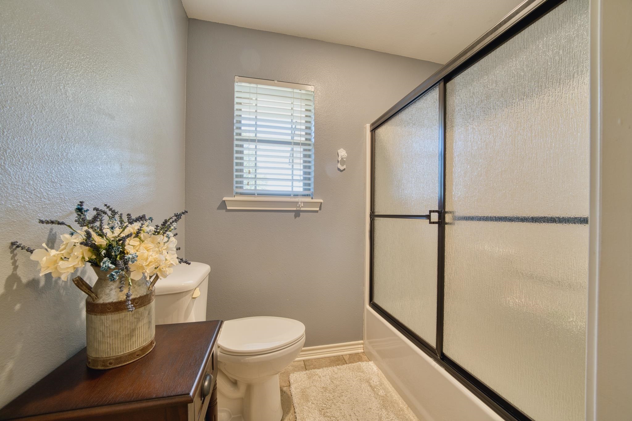 Bathroom with a glass shower enclosure, window with blinds, and flower arrangement on a dark wood vanity.