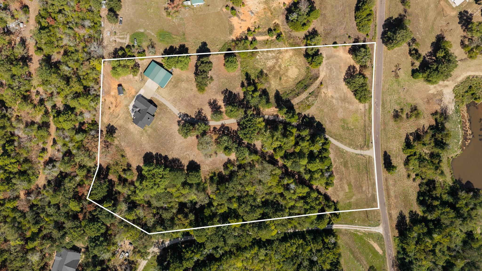 Aerial view of a rural property outlined in white, with a house, green-roofed outbuilding, trees, and road frontage.