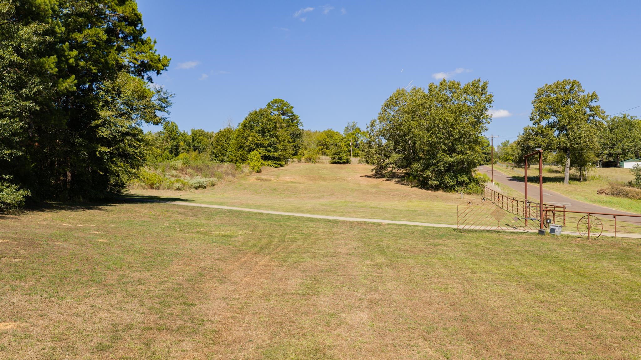 Open grassy property with mature trees, a driveway, and a metal gate beside the road.