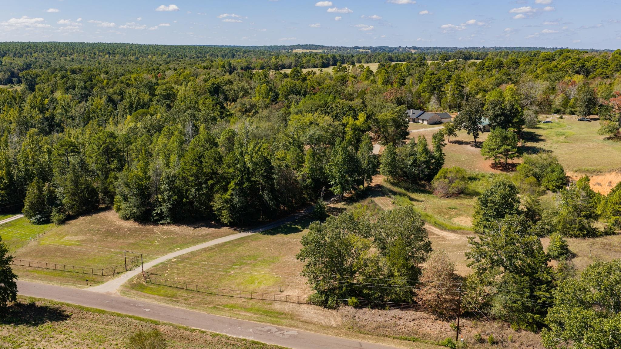 Aerial view of a wooded rural property with a house, open lawns, fencing, and a paved road.