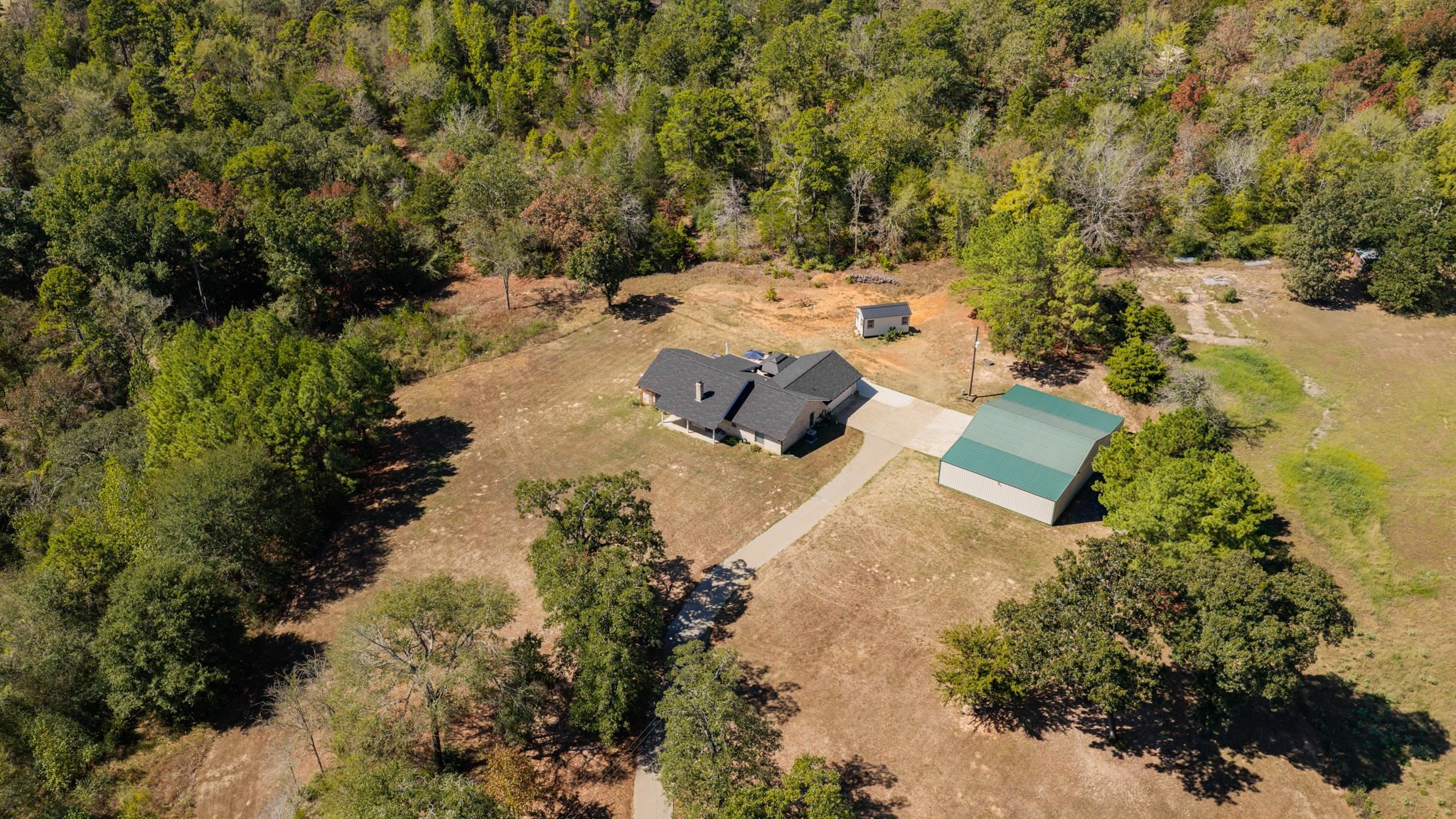 Aerial view of a rural home and green-roofed outbuilding surrounded by wooded acreage.