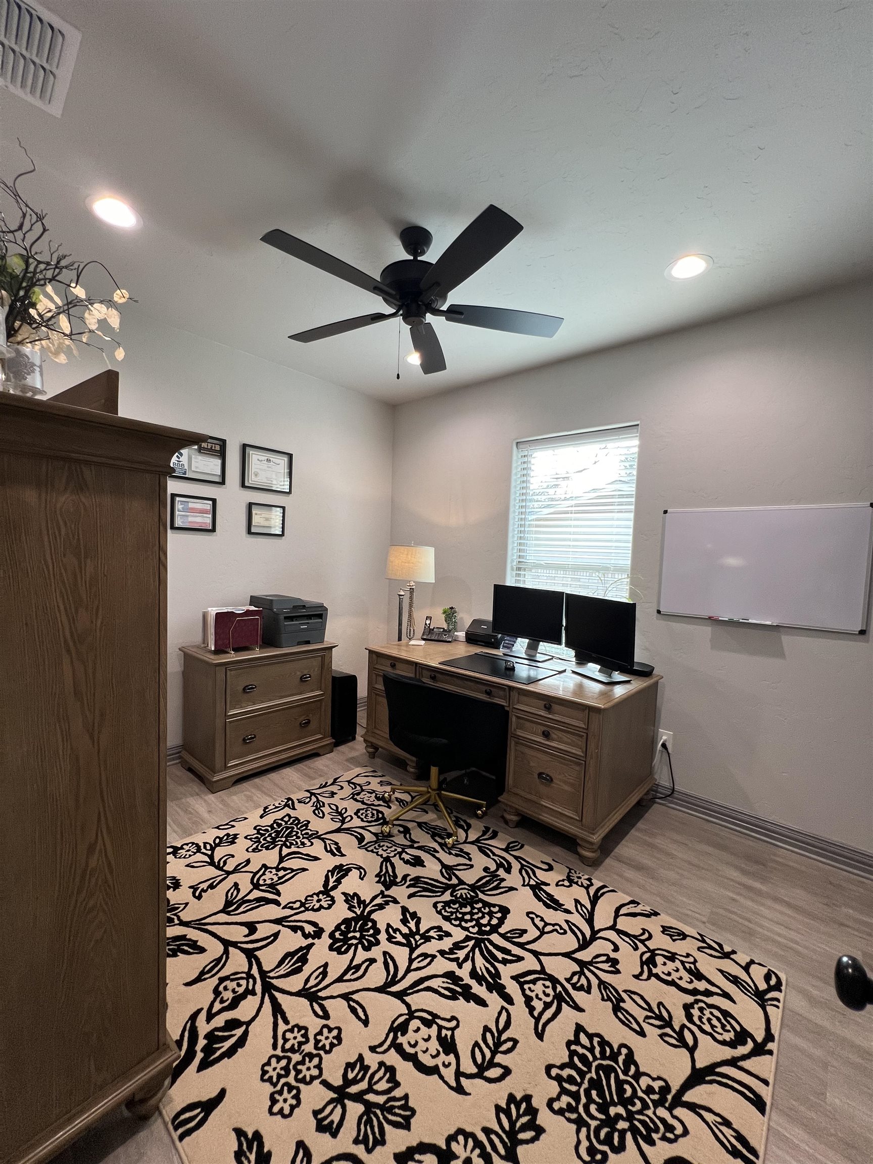 Furnished home office with a wooden desk, dual monitors, and a large black floral rug.