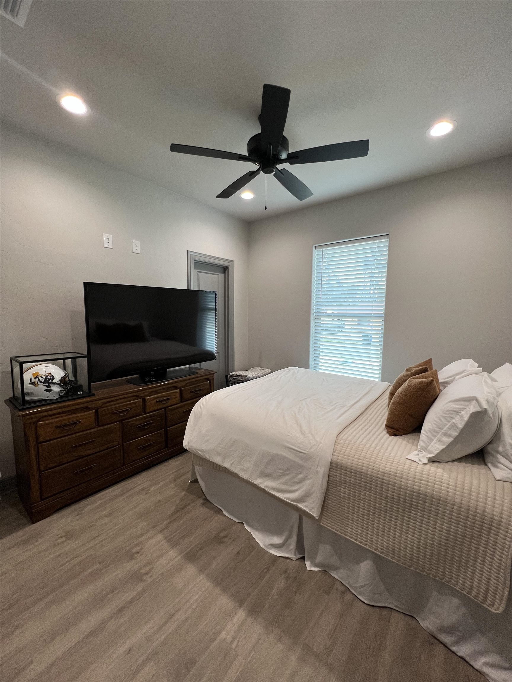 Bedroom with white bedding, brown accent pillows, a television on a dark wood dresser, and a ceiling fan.