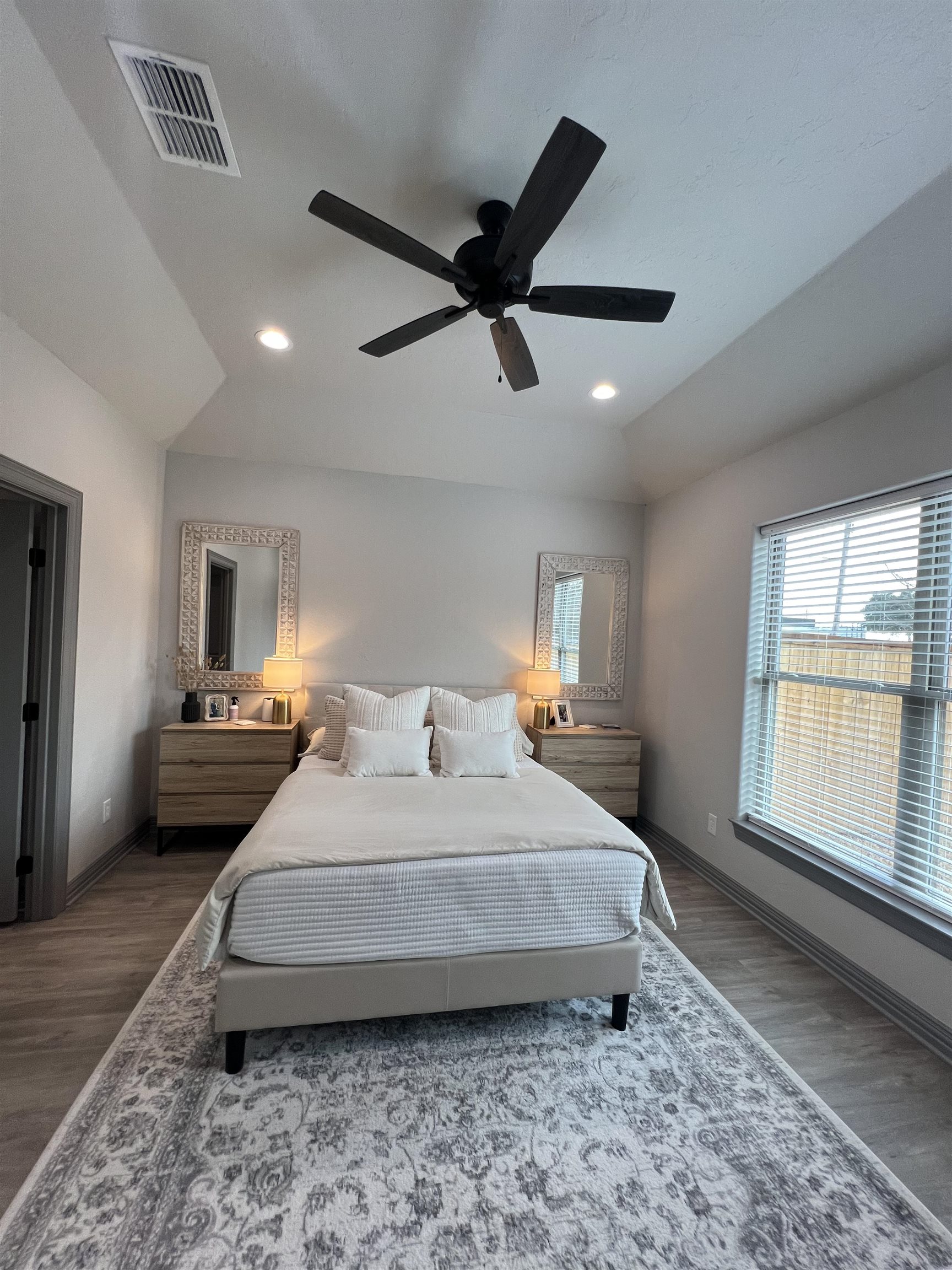 Furnished bedroom with a white bed, matching nightstands, vaulted ceiling, and large window with blinds.