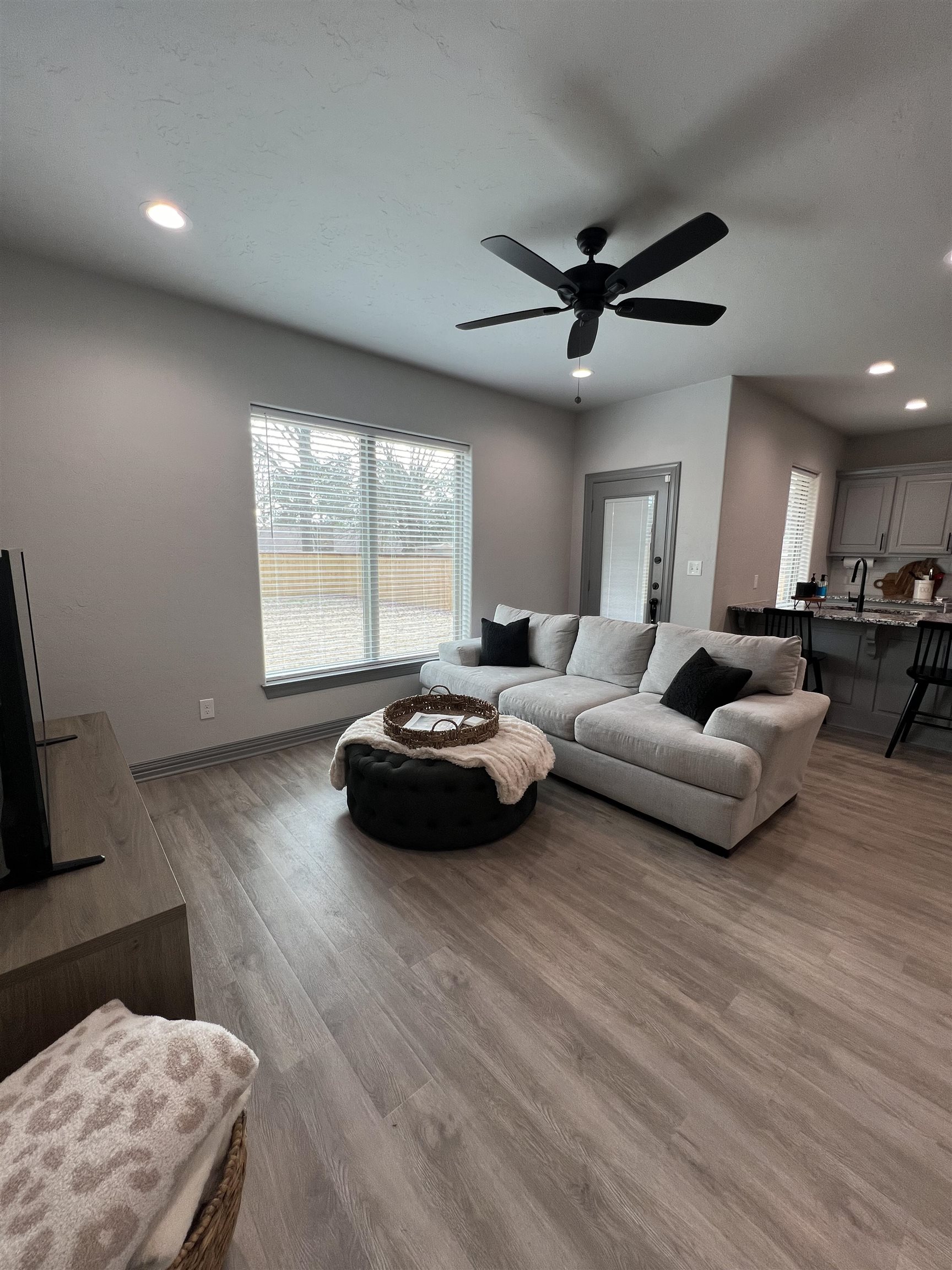 Living room with a light sectional, black ceiling fan, large window, and open kitchen.