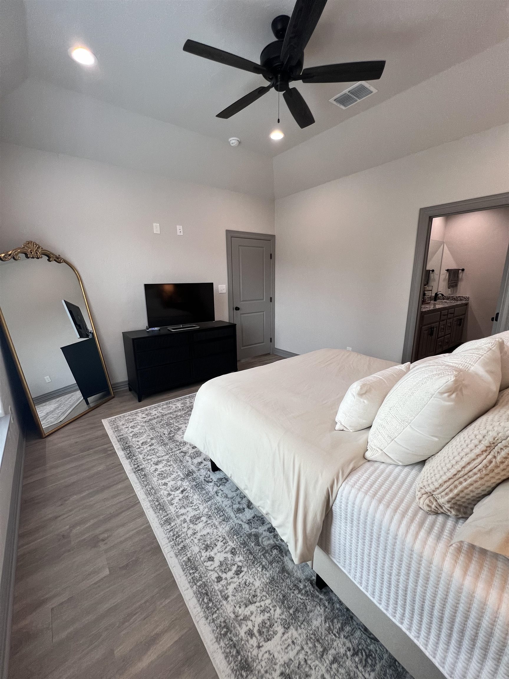 Bedroom with a white bed, patterned rug, black dresser, and ornate floor mirror.