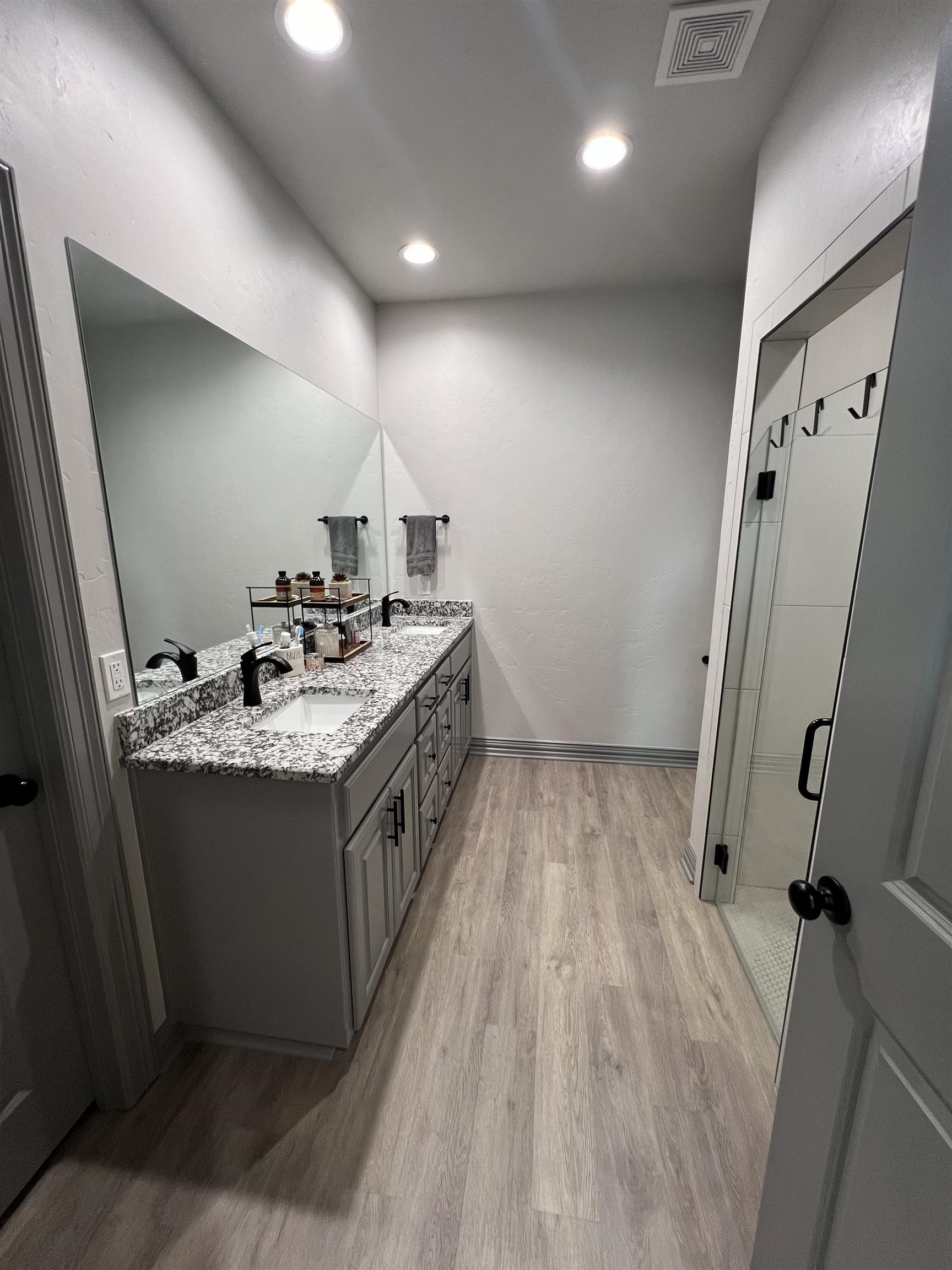 Bathroom with a double vanity, speckled countertop, large mirror, and glass-enclosed shower.