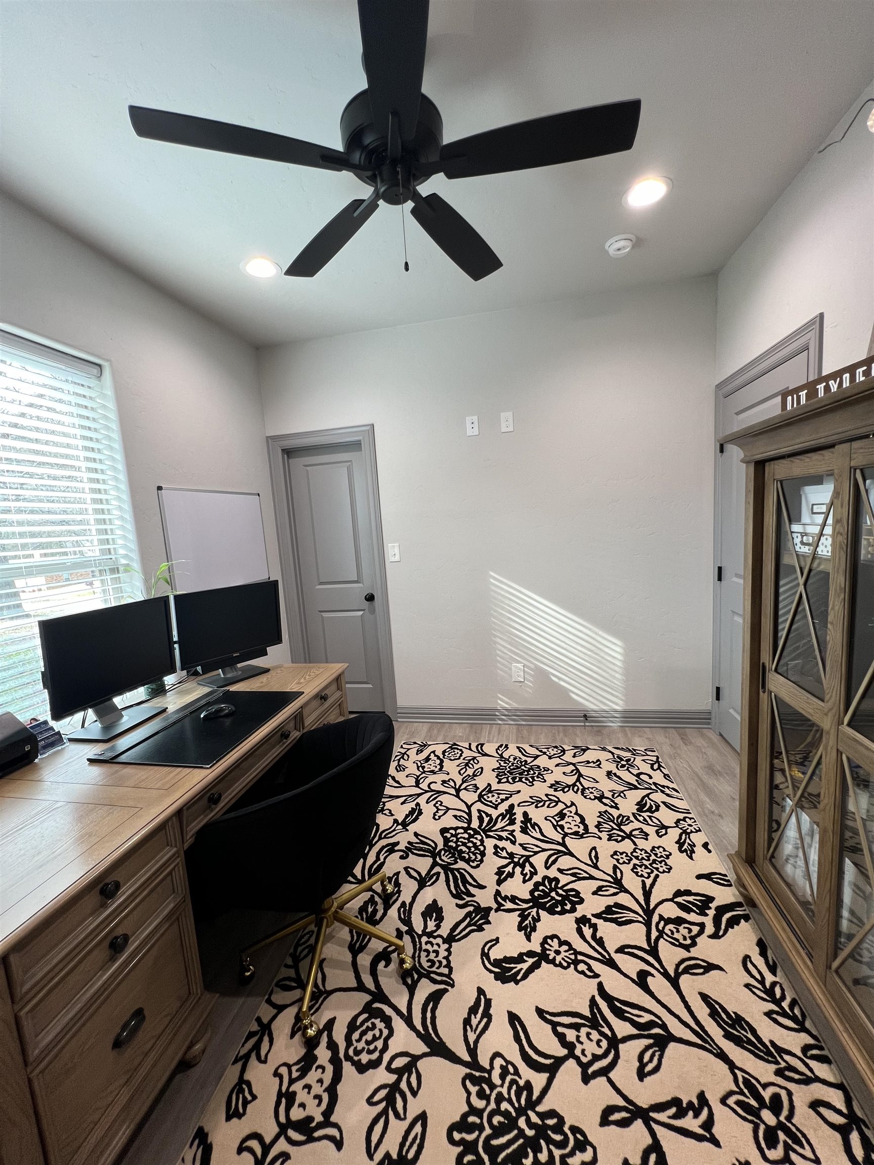 Home office with a wooden desk, dual monitors, patterned rug, and glass-front cabinet