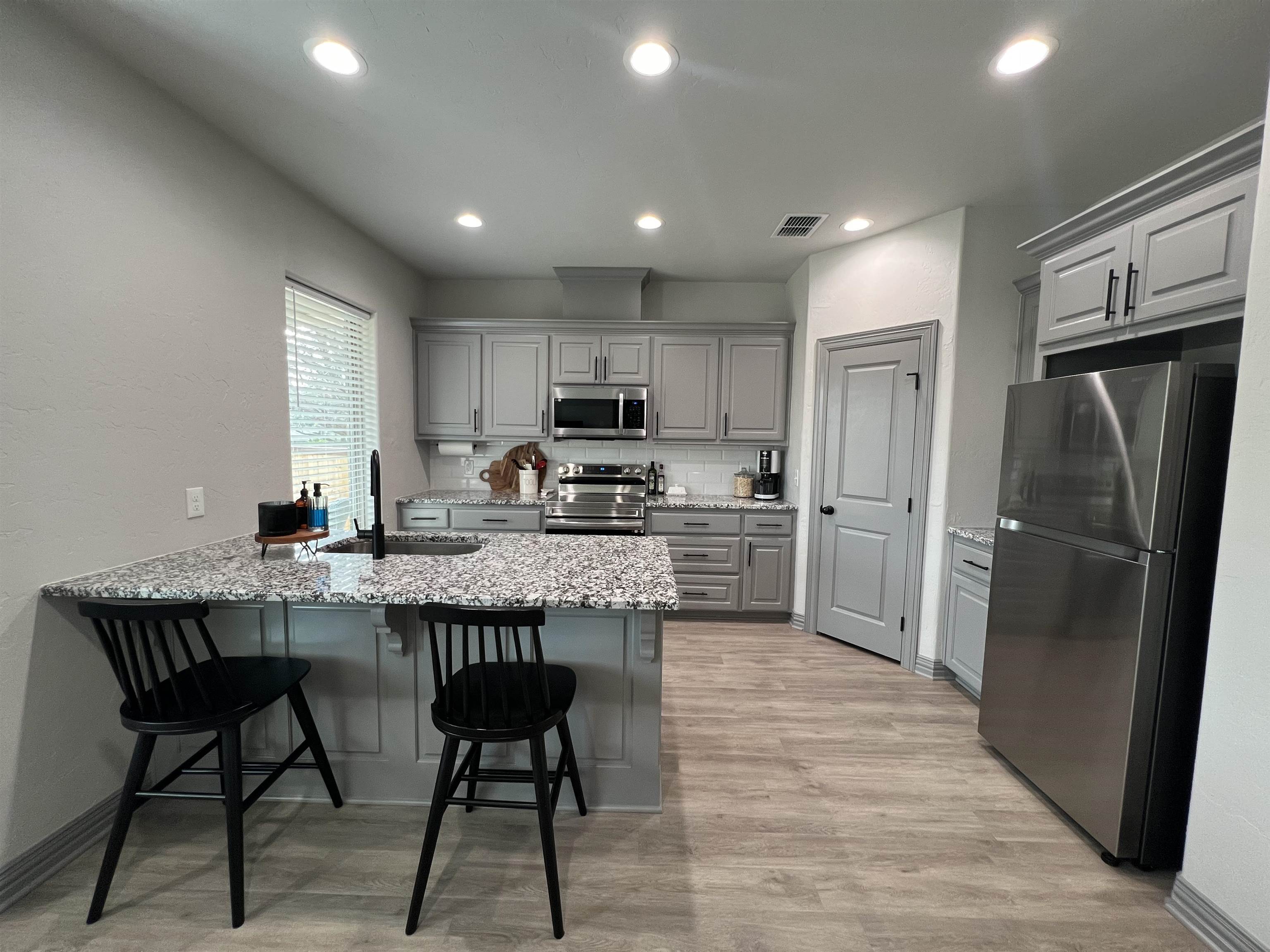 Kitchen with gray cabinets, stainless steel appliances, and a granite island with sink and two black chairs.
