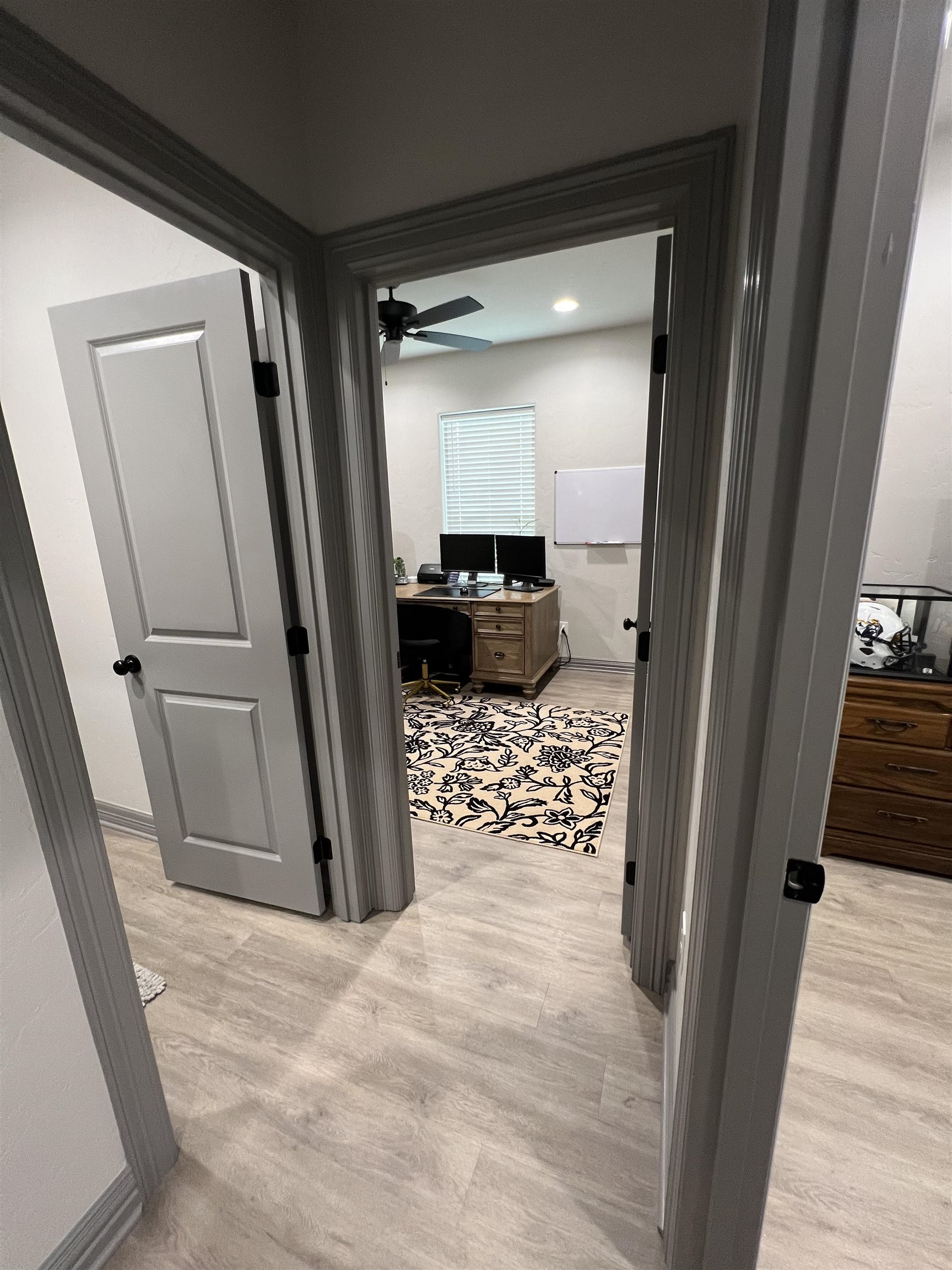 Home office viewed through a doorway, with a wooden desk, dual monitors, ceiling fan, and floral rug.