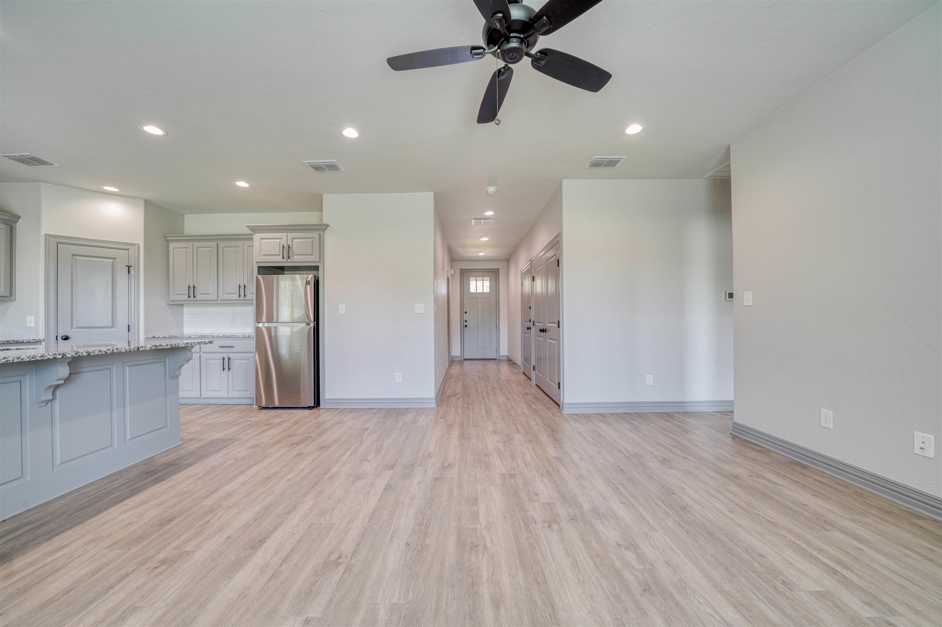 Vacant kitchen and living area with a granite breakfast bar, stainless refrigerator, and hallway to the front door.