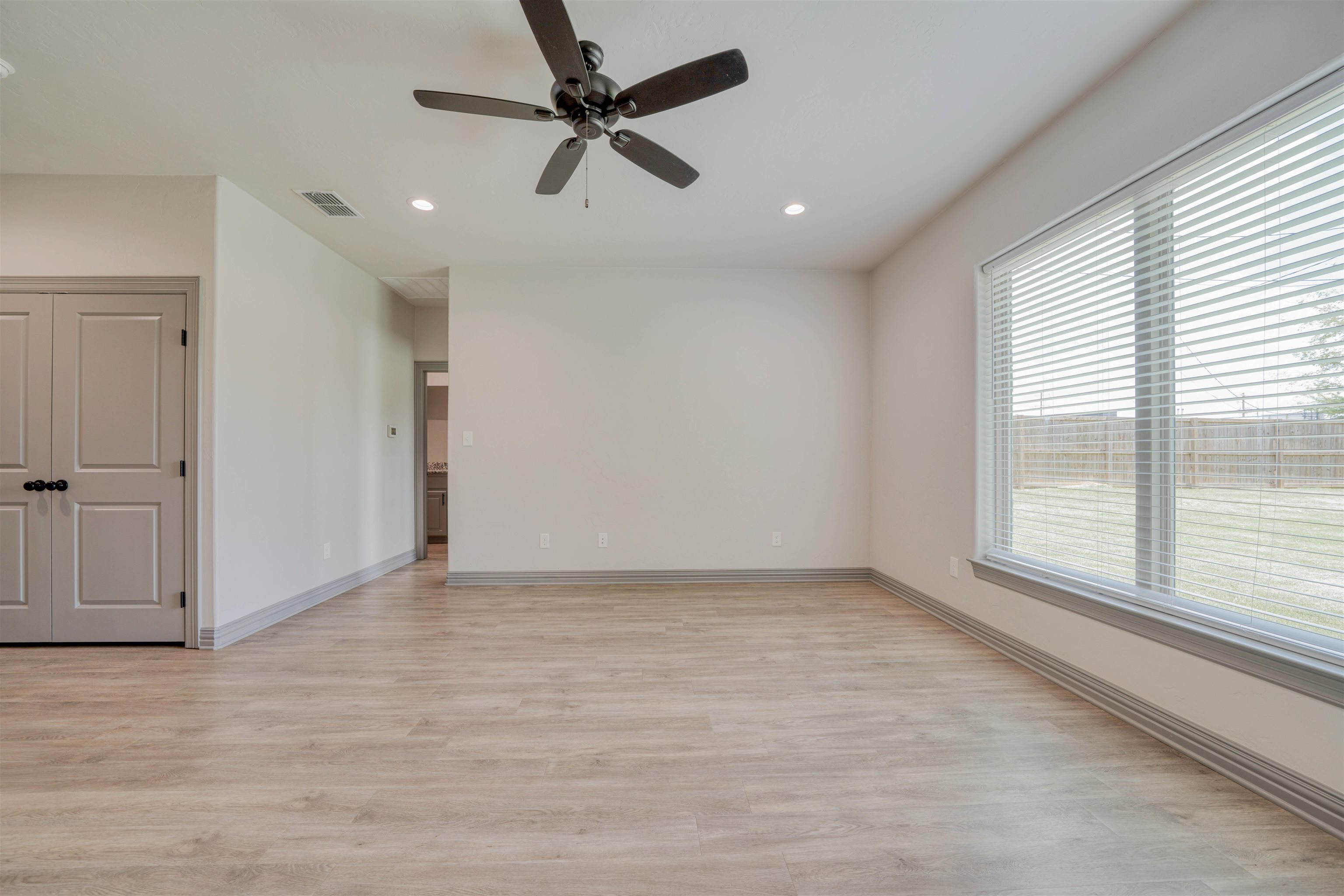 Empty room with wood-look flooring, ceiling fan, and a large window overlooking a fenced yard.