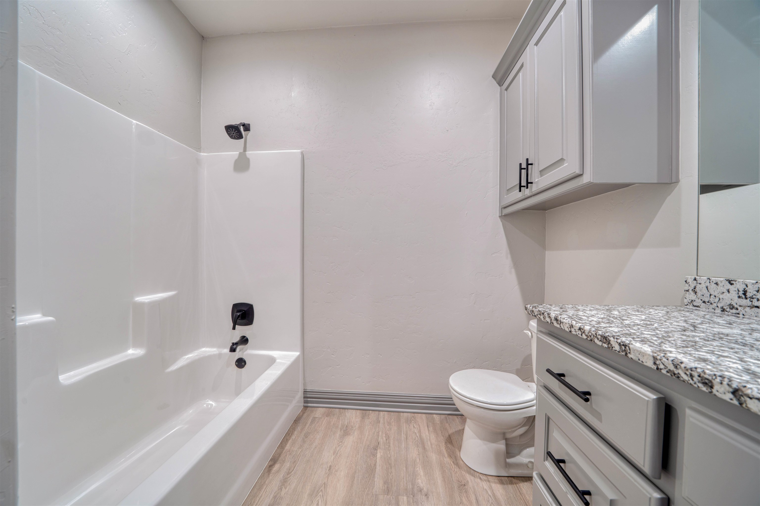 Bathroom with a white tub and shower beside a toilet and gray vanity with granite countertop.
