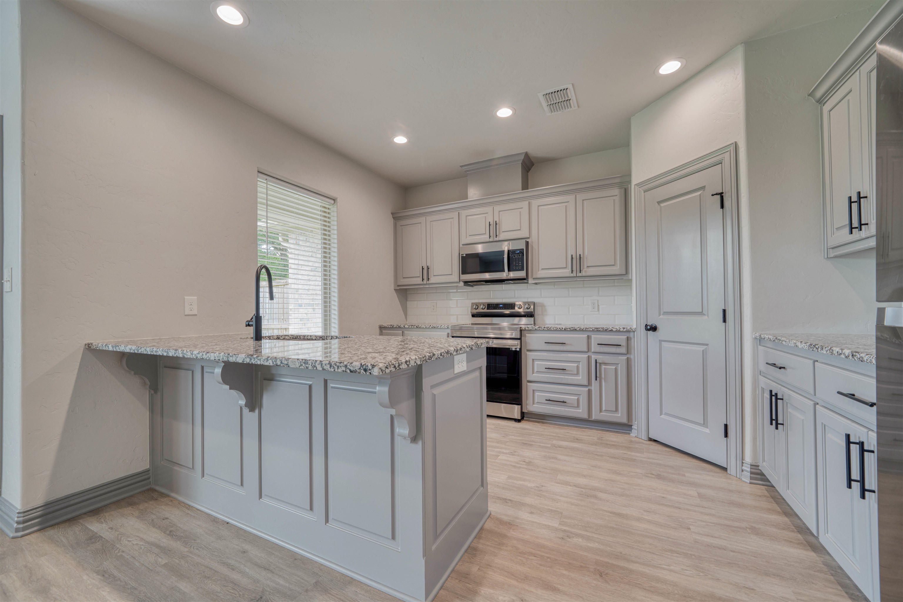 Kitchen with white cabinets, granite island, stainless steel appliances, and light wood-look flooring.