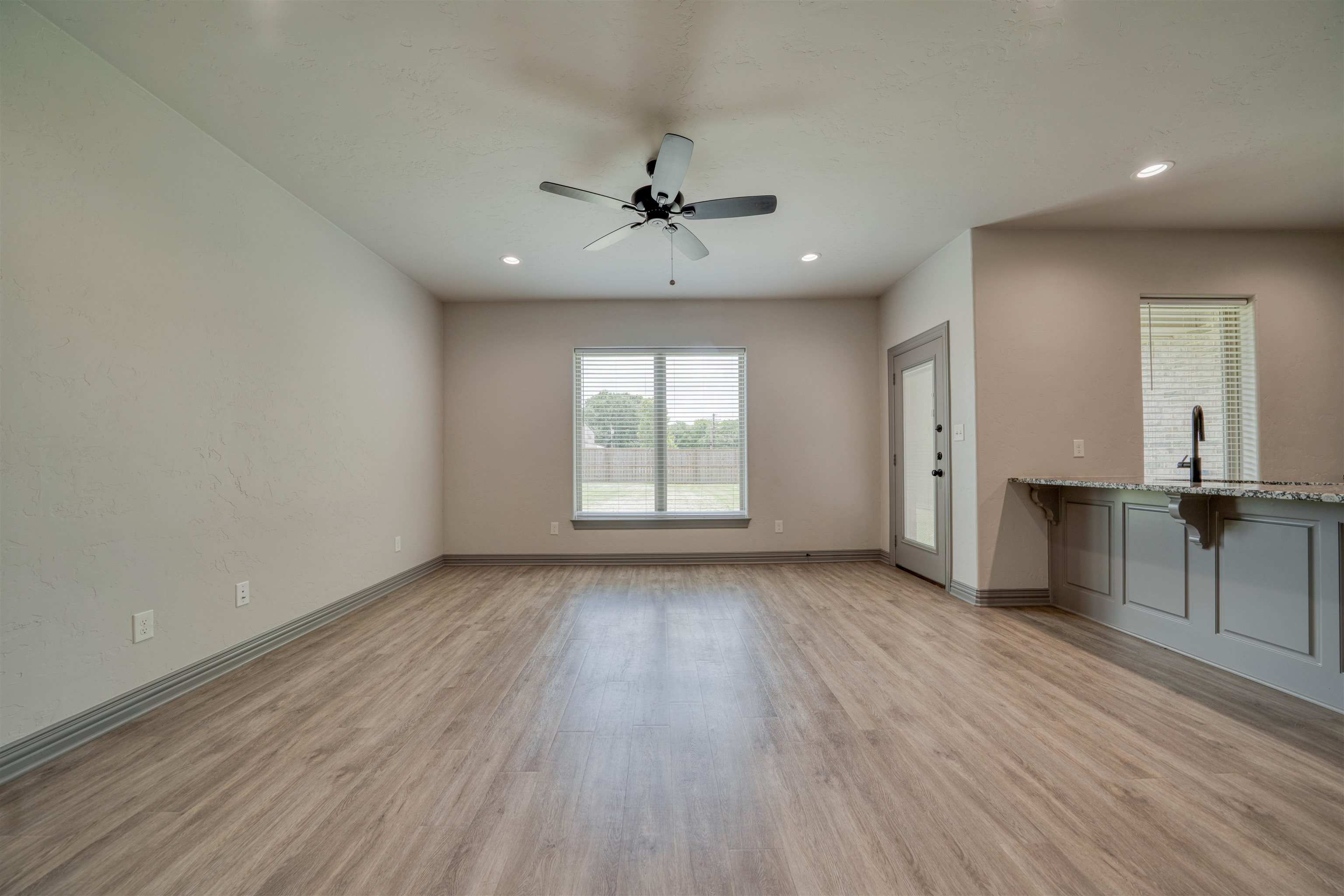 Vacant living area with wood-look floors, ceiling fan, window, and gray kitchen peninsula.