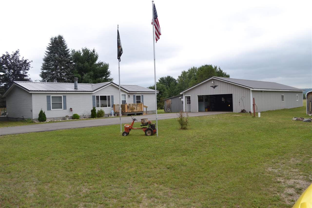 Gray single-story home and large detached metal garage on a grassy lot, with two flagpoles out front.