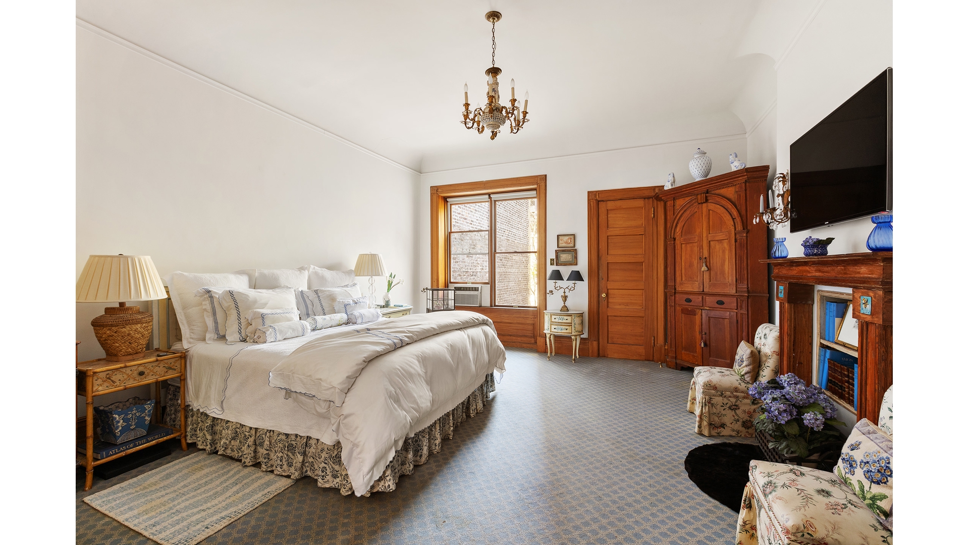 Furnished bedroom with a large white bed, ornate wooden wardrobe, and fireplace.