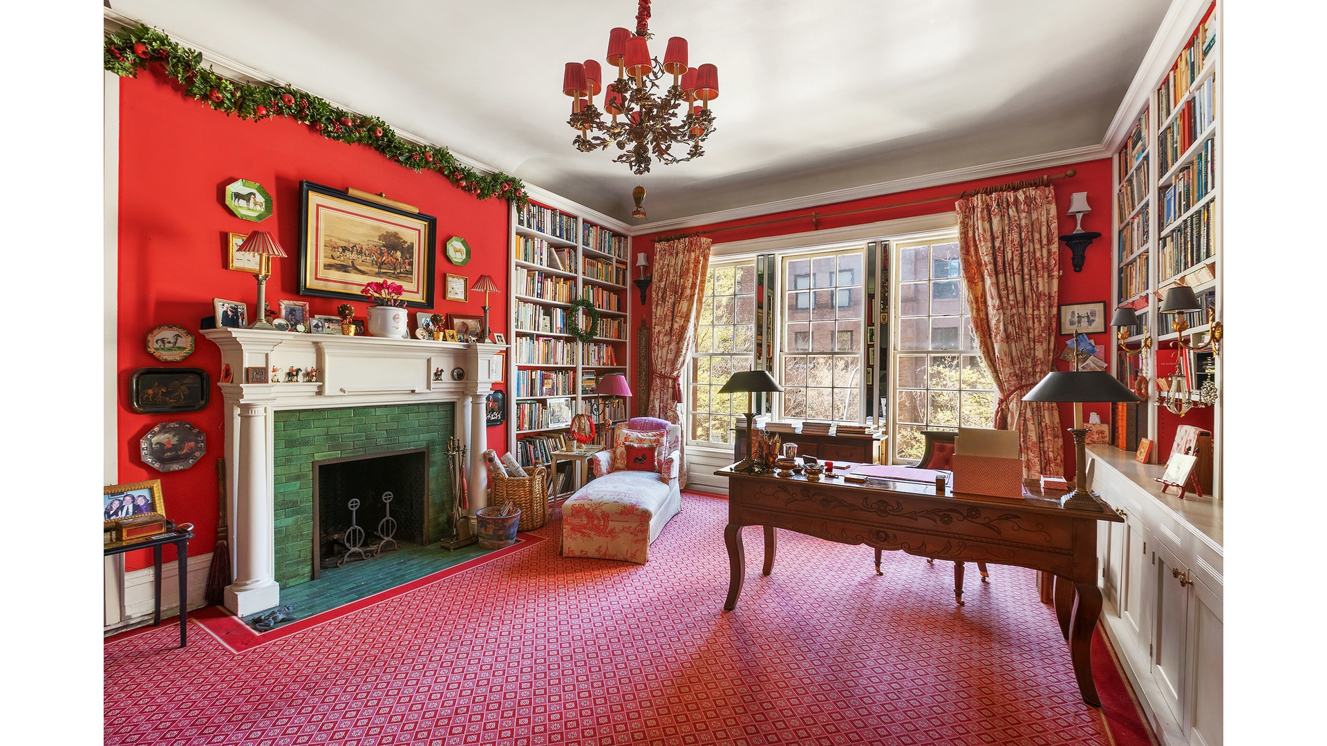 Red-walled study with bookshelves, a green-tiled fireplace, and a carved wooden desk by tall windows.