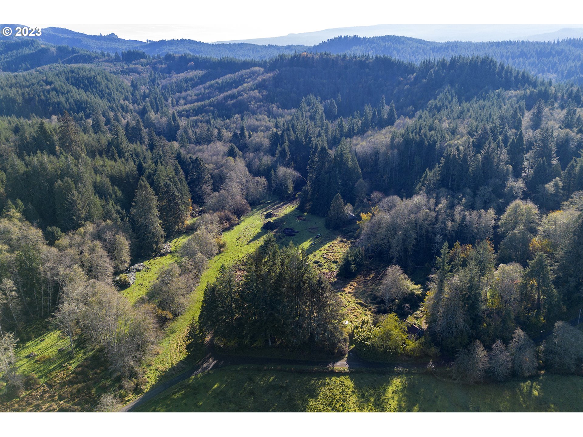 Aerial view of a grassy clearing winding through dense forest, with a road and wooded hills beyond.