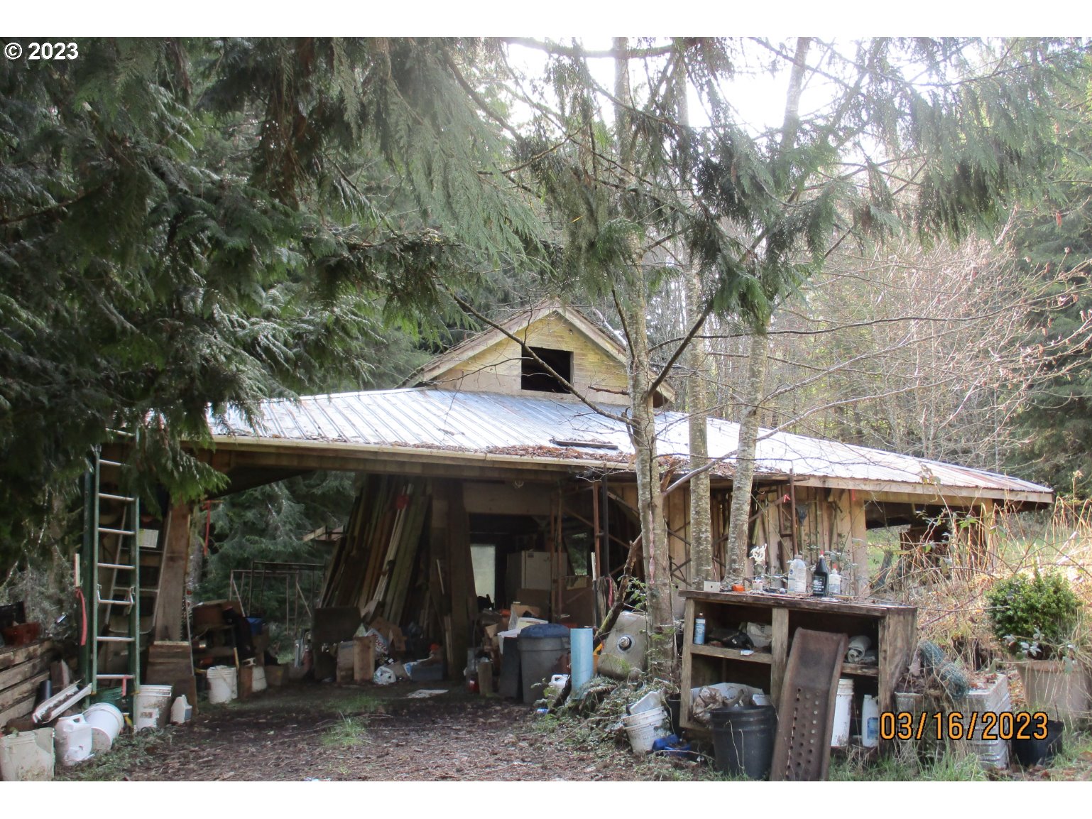 Open-sided workshop with a metal roof, surrounded by trees and filled with tools and stored materials.