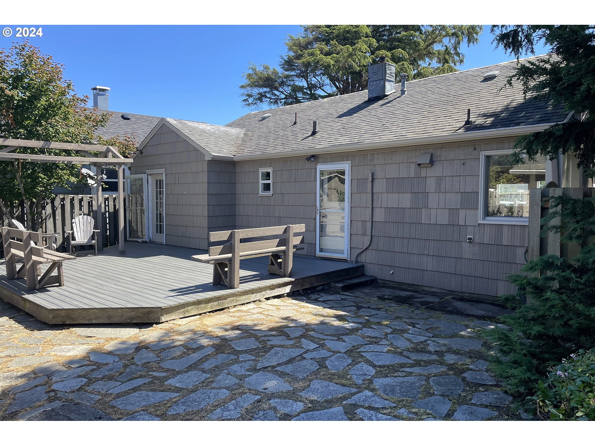 Rear of a gray shingle-sided house with a wooden deck, outdoor seating, and a stone patio.