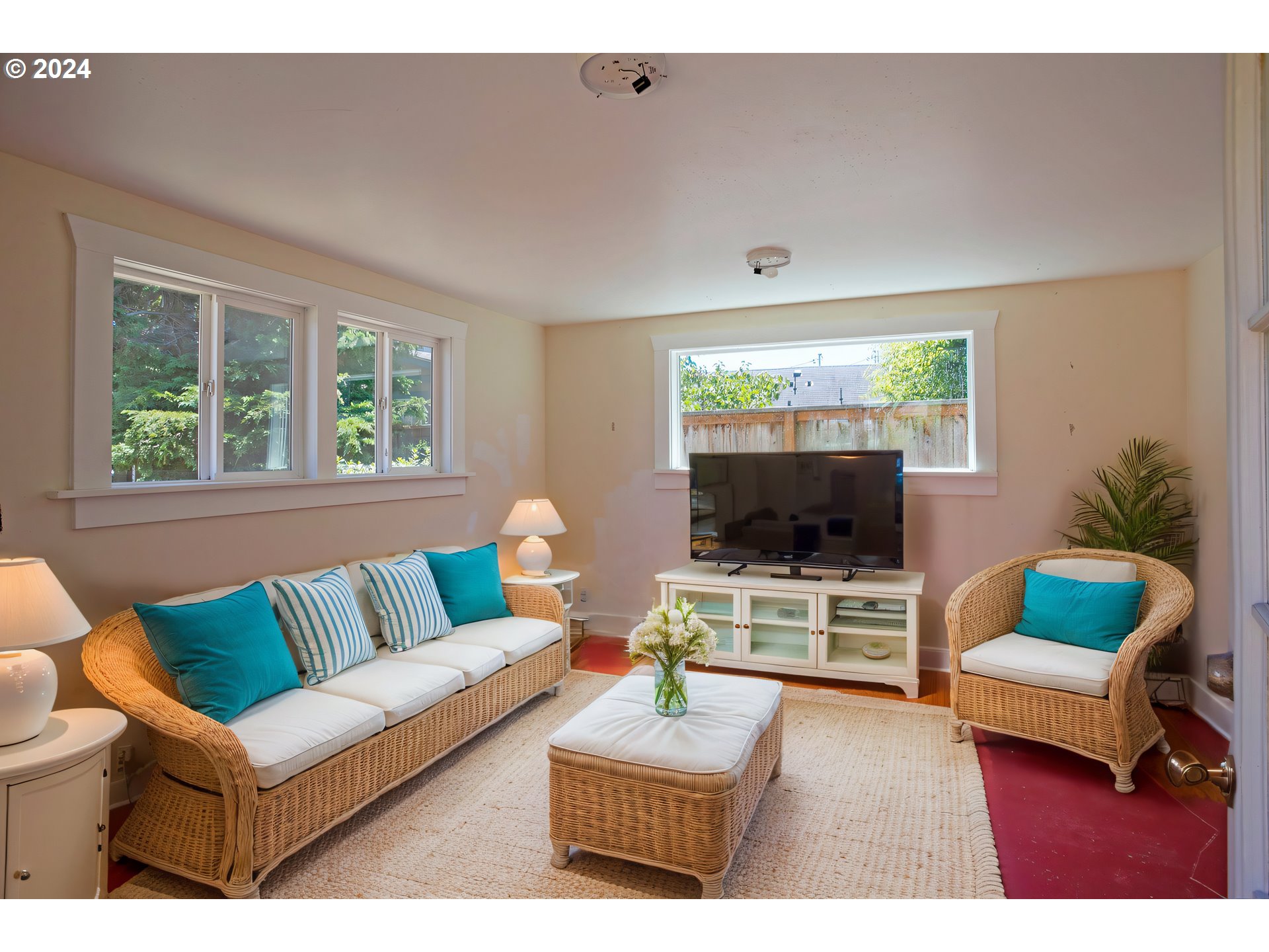 Living room with wicker seating, turquoise pillows, a TV console, and large windows overlooking a fenced yard.