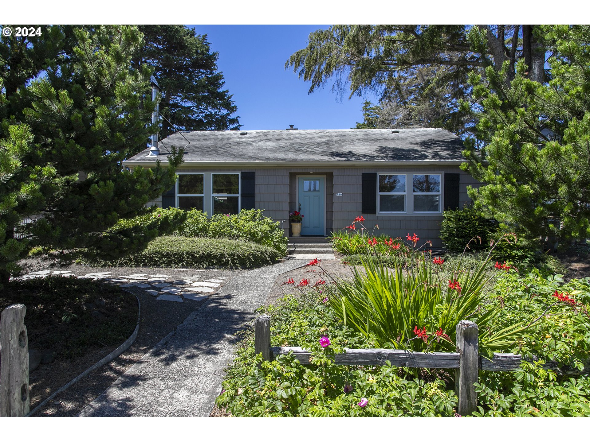Gray single-story home with a blue front door, mature trees, and a colorful garden along the entry path.