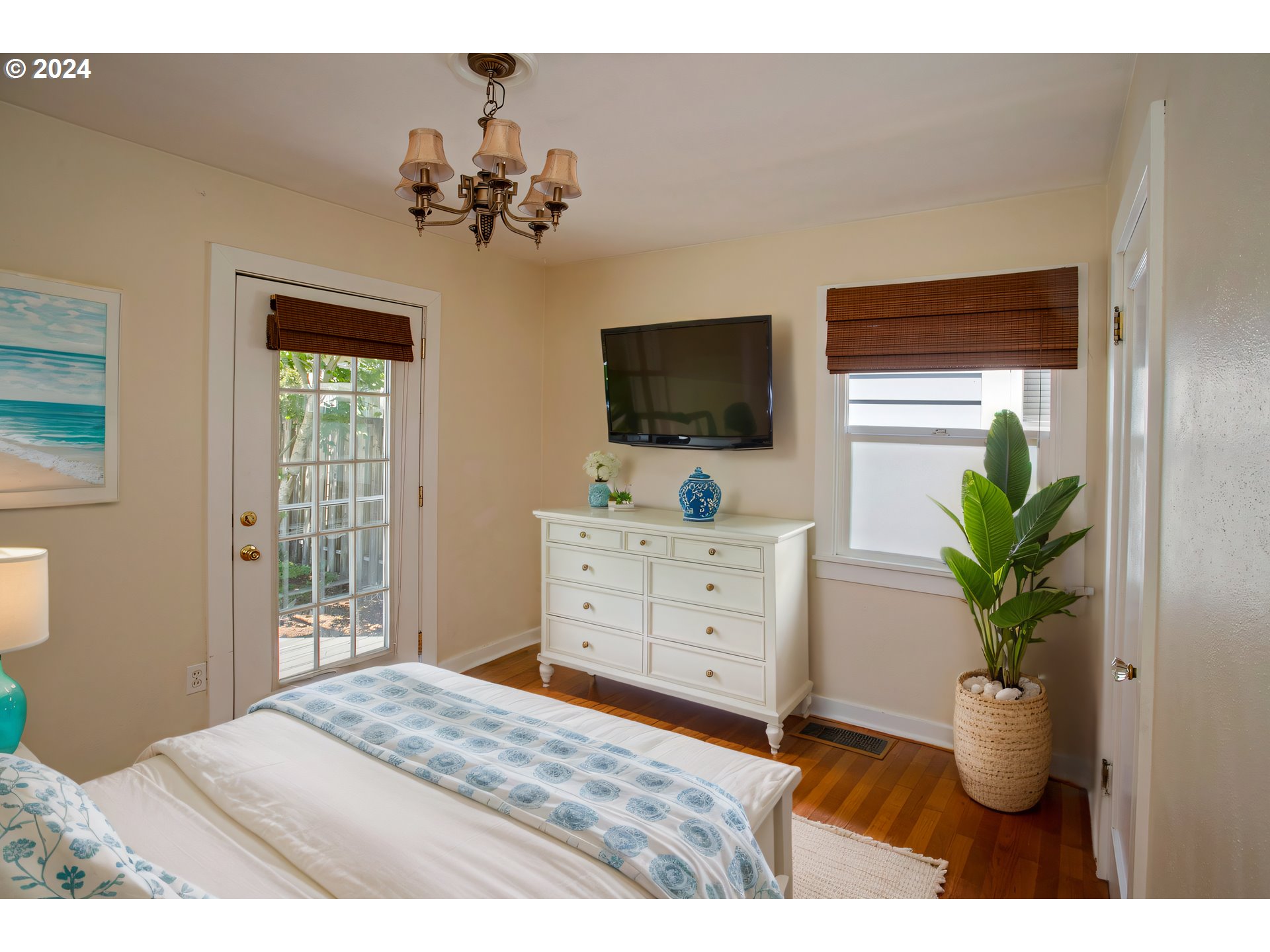 Bedroom with blue-and-white bedding, a white dresser, wall-mounted TV, and potted plant by the window.