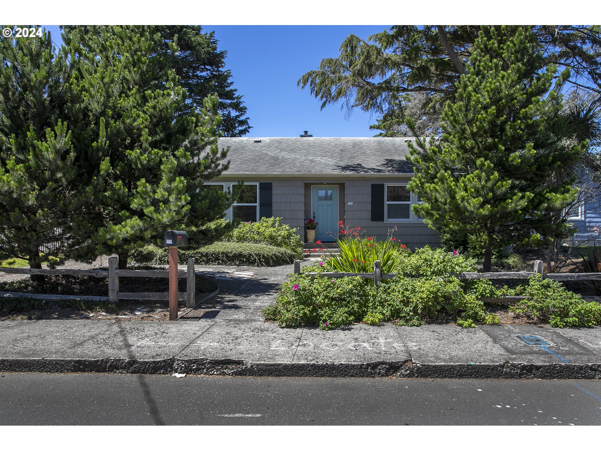 Gray single-story house with a blue front door, flowering garden, and tall evergreens.