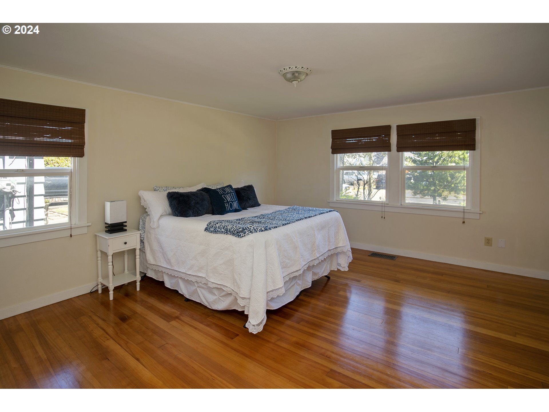 Furnished bedroom with a white bed, dark blue pillows, three windows, and polished hardwood floors.