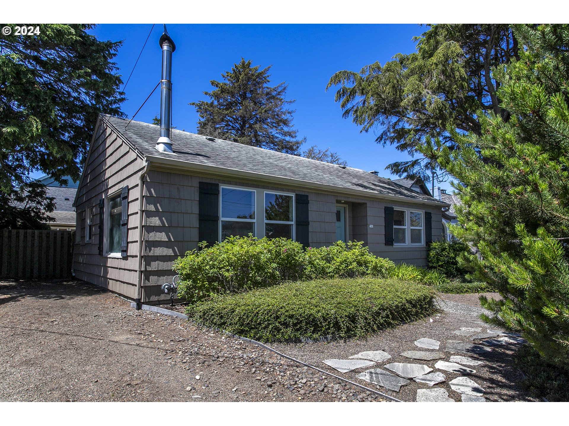 Gray shingle-sided single-story house with dark shutters, front shrubs, and a gravel yard with flagstone path.