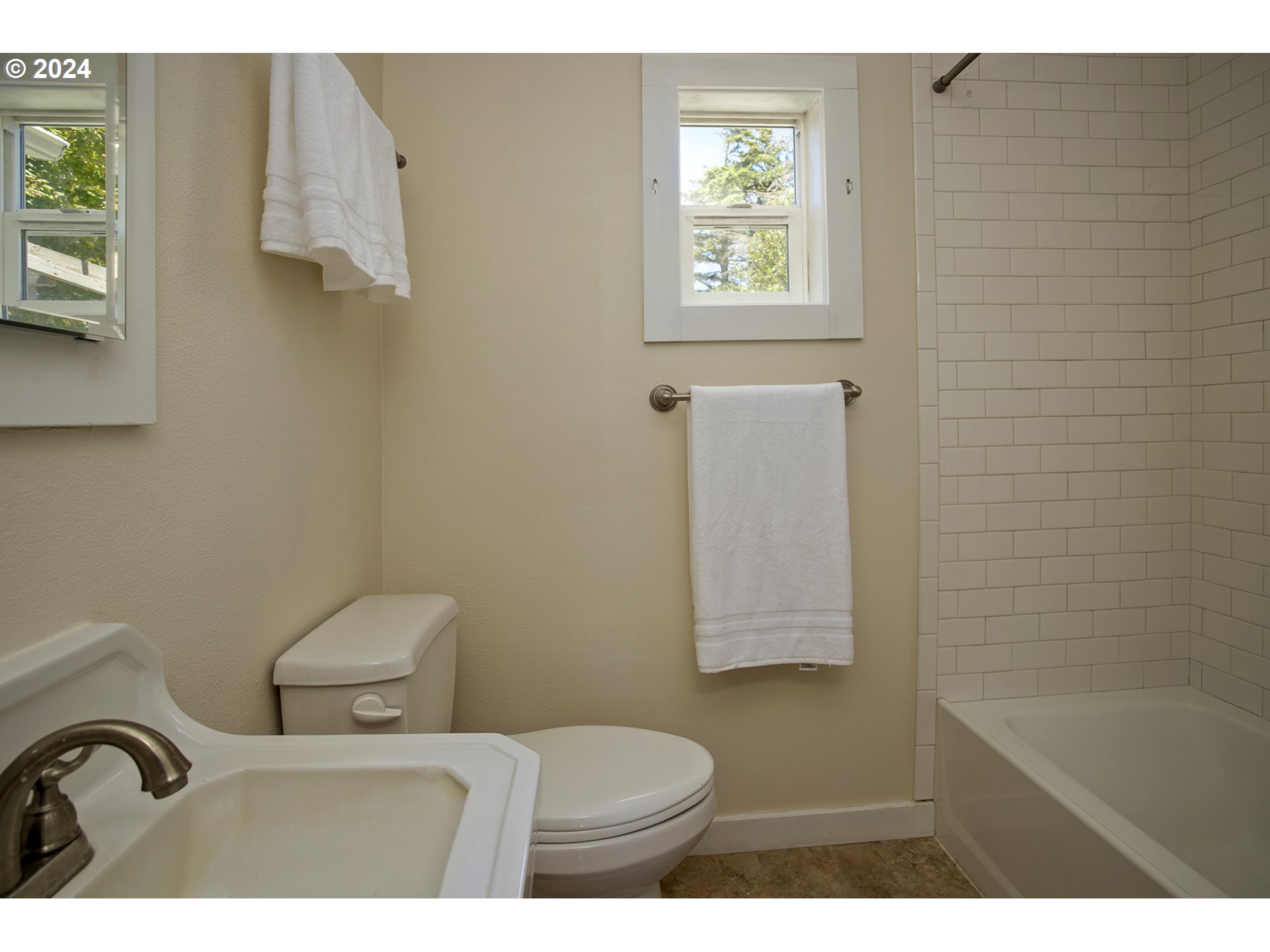 Bathroom with a subway-tile tub surround, two windows, white towels, sink, and toilet.