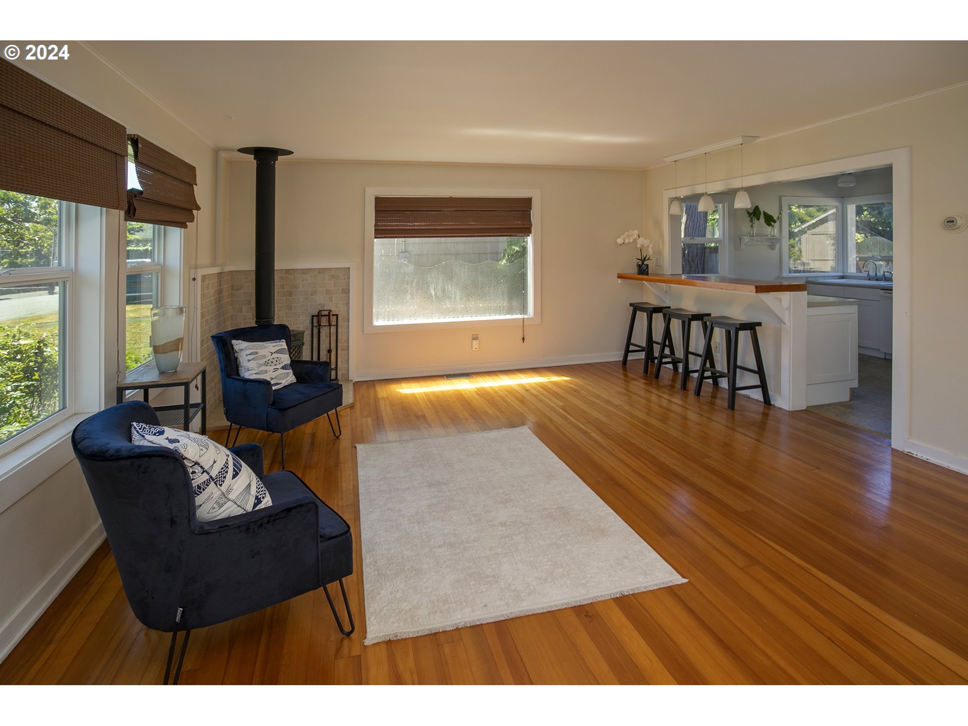 Furnished living room with hardwood floors, navy armchairs, and a wood stove beside the kitchen bar.