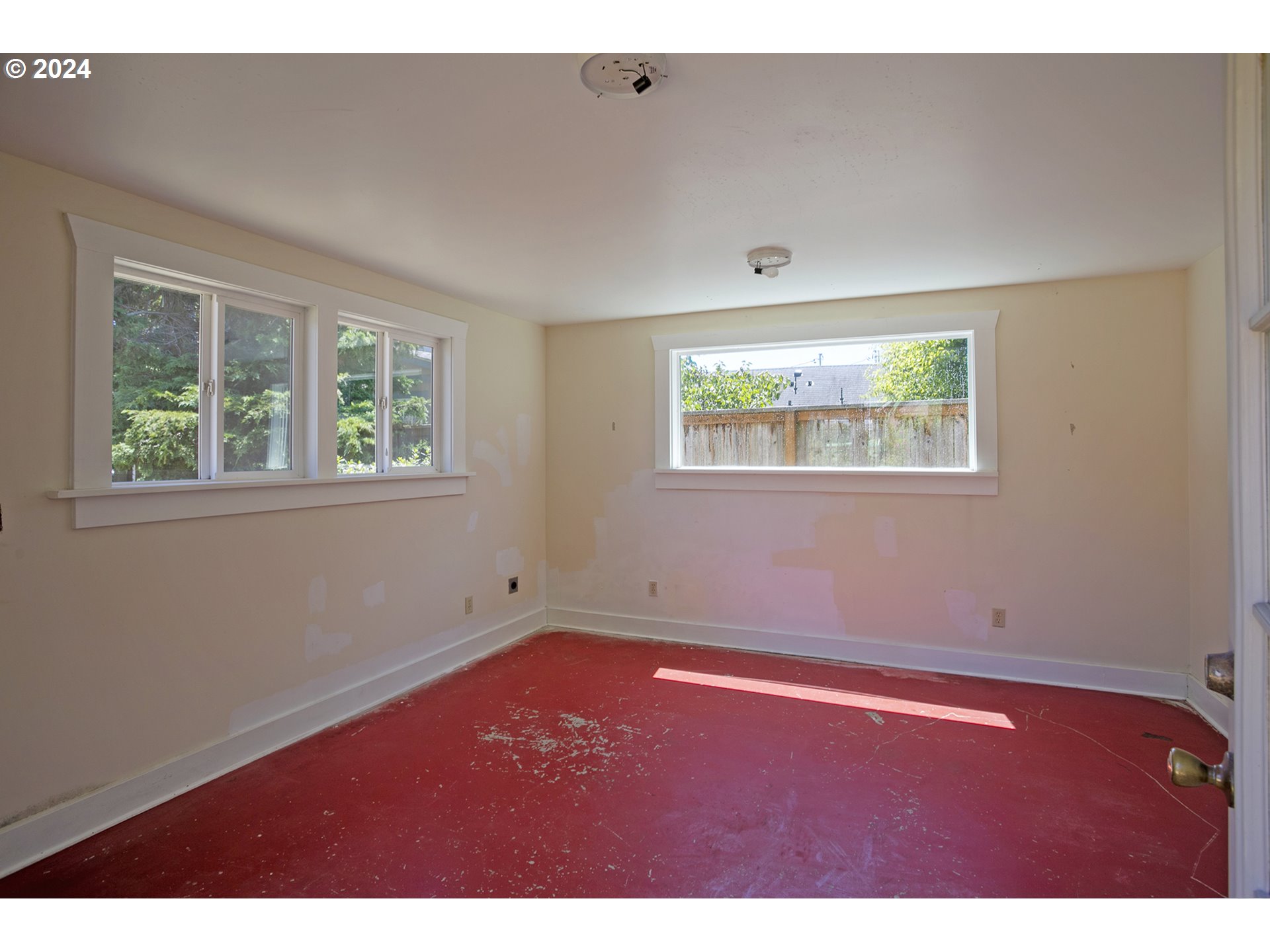 Empty room with patched cream walls, a worn red floor, and several white-trimmed windows.