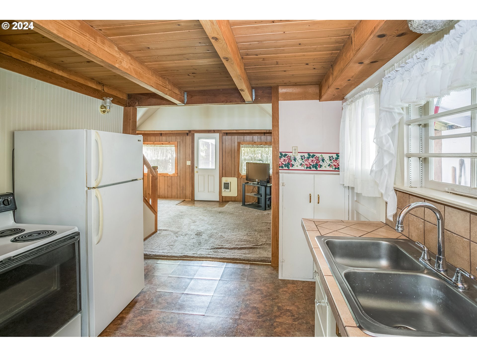 Kitchen with exposed wood ceiling beams, a double sink, and an adjoining carpeted room.